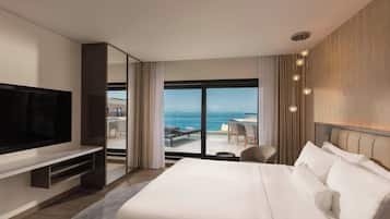 Una camera d'albergo moderna con un letto grande, una TV a schermo piatto, un balcone con vista sull'oceano e un'area salotto con tavolo e sedie.