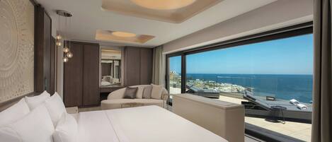 A modern hotel room with a large bed, a sitting area, and a balcony with a view of the sea.