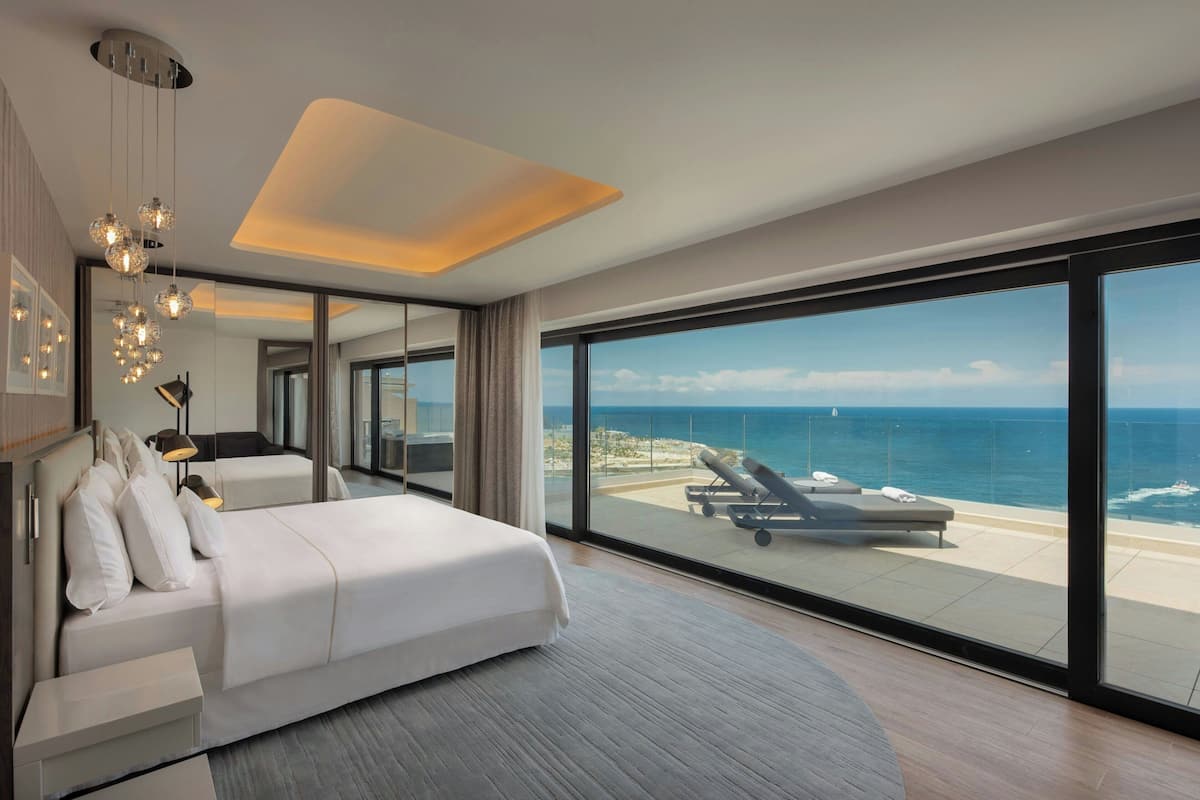 A modern hotel room with a large bed, a seating area with a view of the ocean, and a balcony with sun loungers.