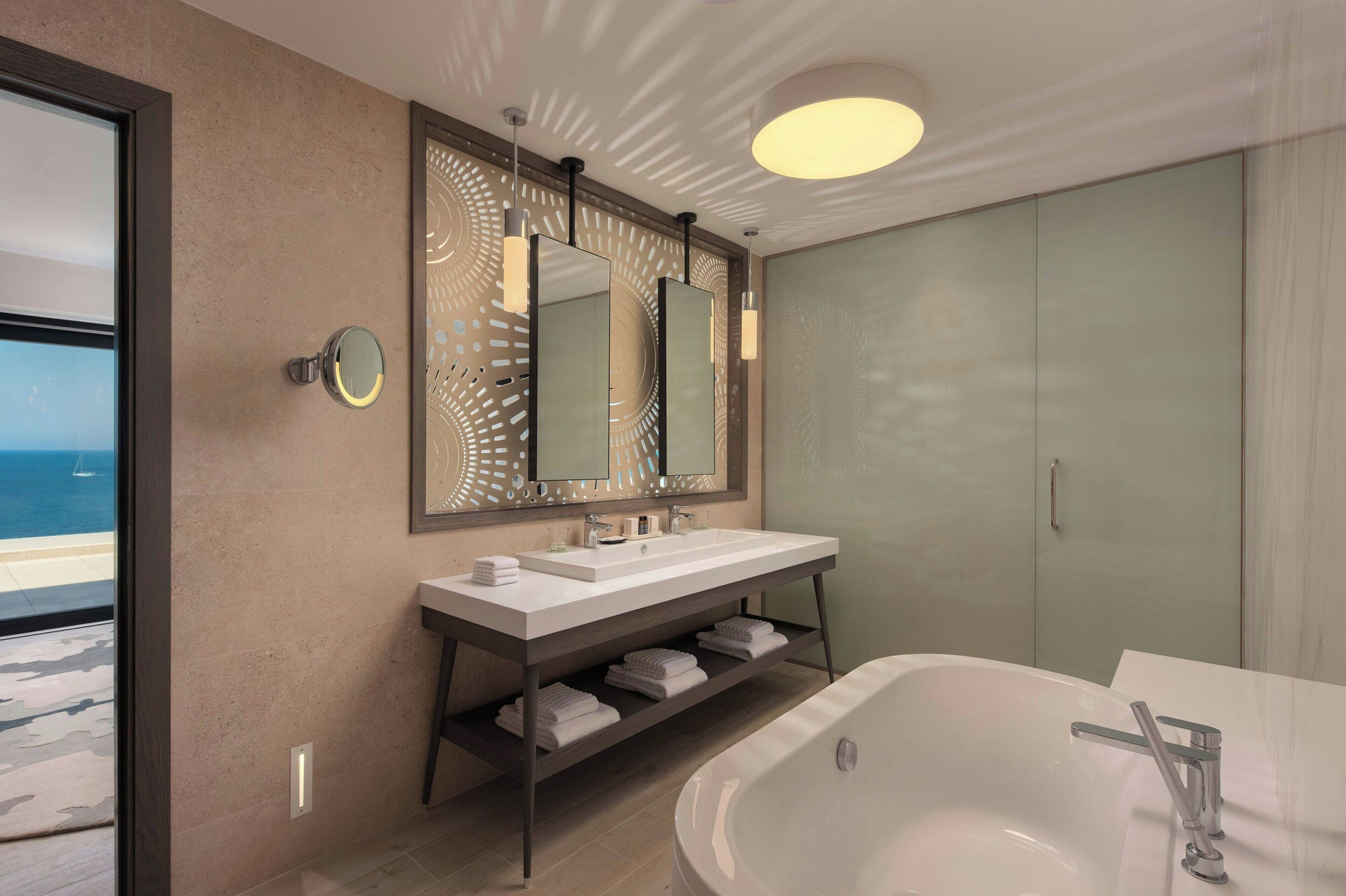 A modern bathroom with a large bathtub, a double vanity with a mirror, and a view of the sea.
