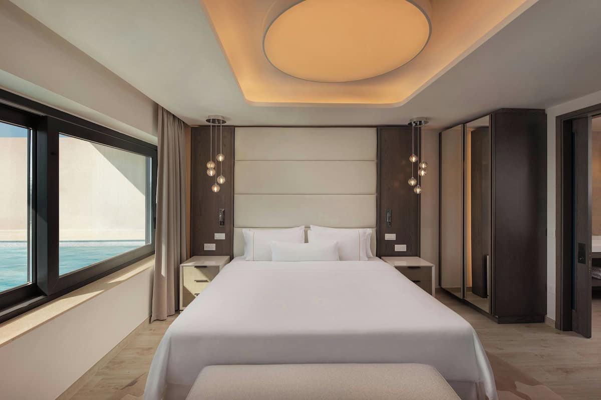 A modern hotel room with a large bed, bedside tables, a nightstand, a wardrobe, and a view of the ocean.