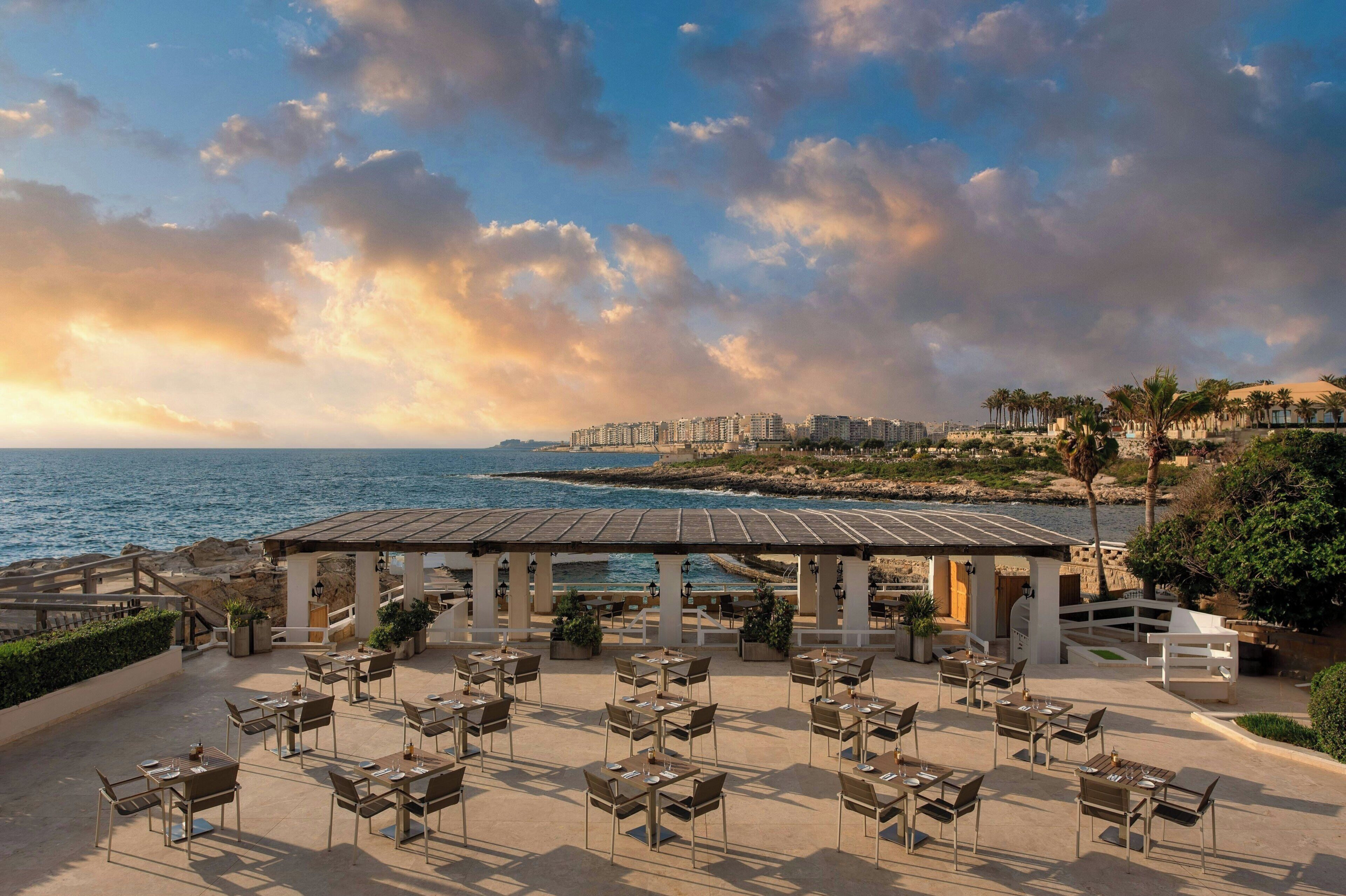 An outdoor dining area with tables and chairs, overlooking the sea and a coastal cityscape.