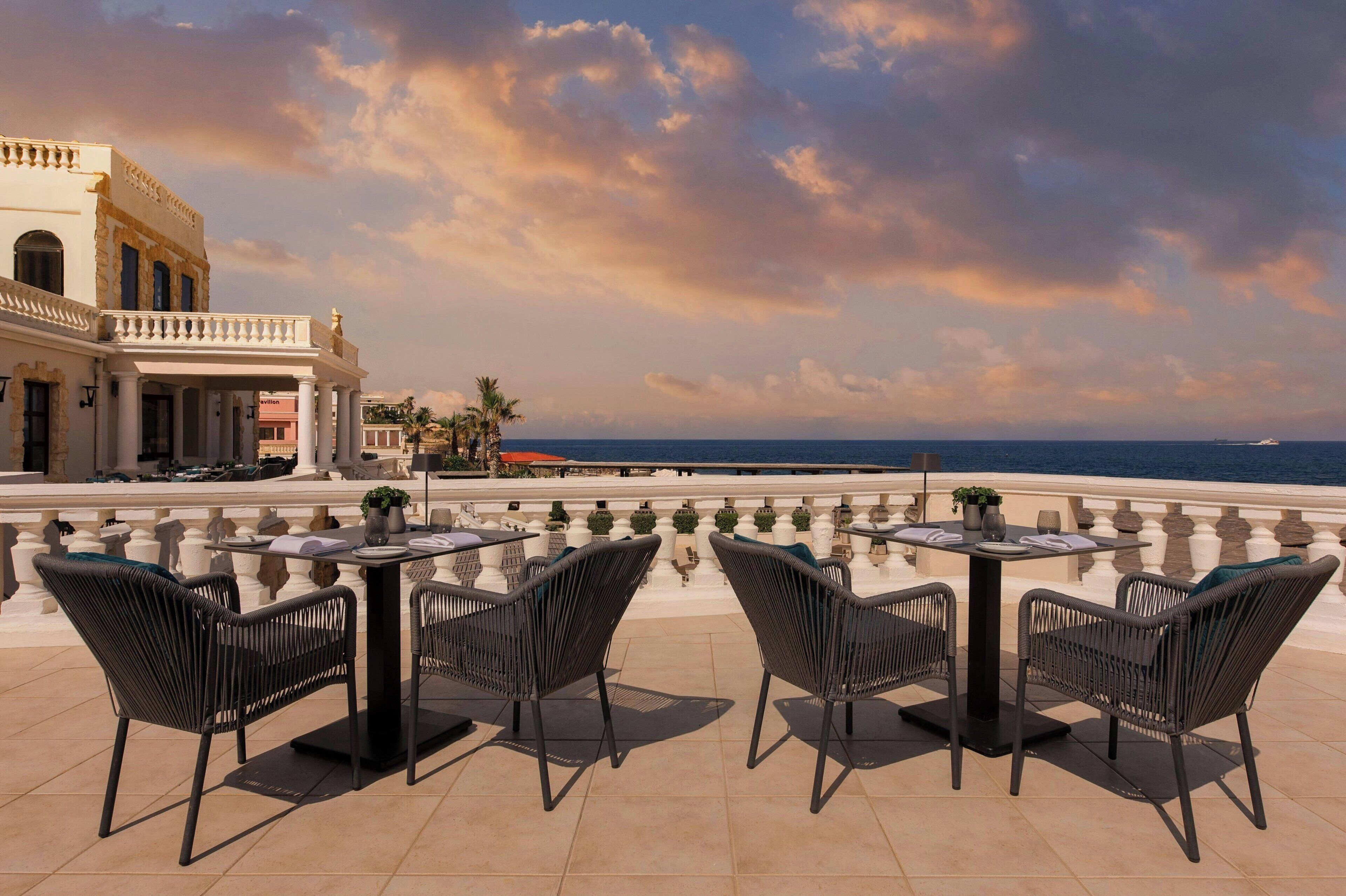 An outdoor terrace with tables and chairs, overlooking a body of water and a building with classical architecture.