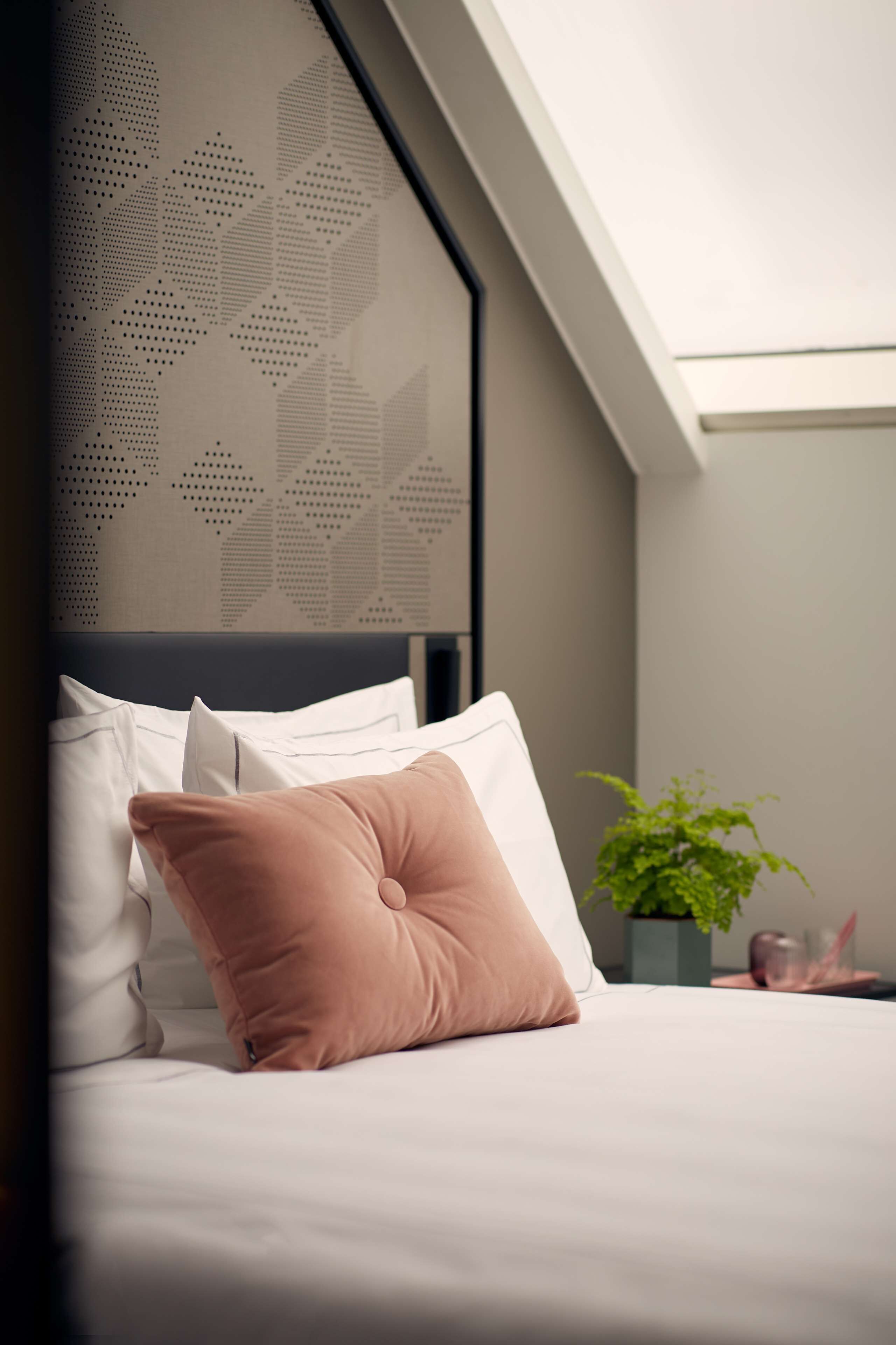 A neatly made bed with white and pink pillows, a decorative wall with a geometric pattern, and a small green plant on a nightstand.