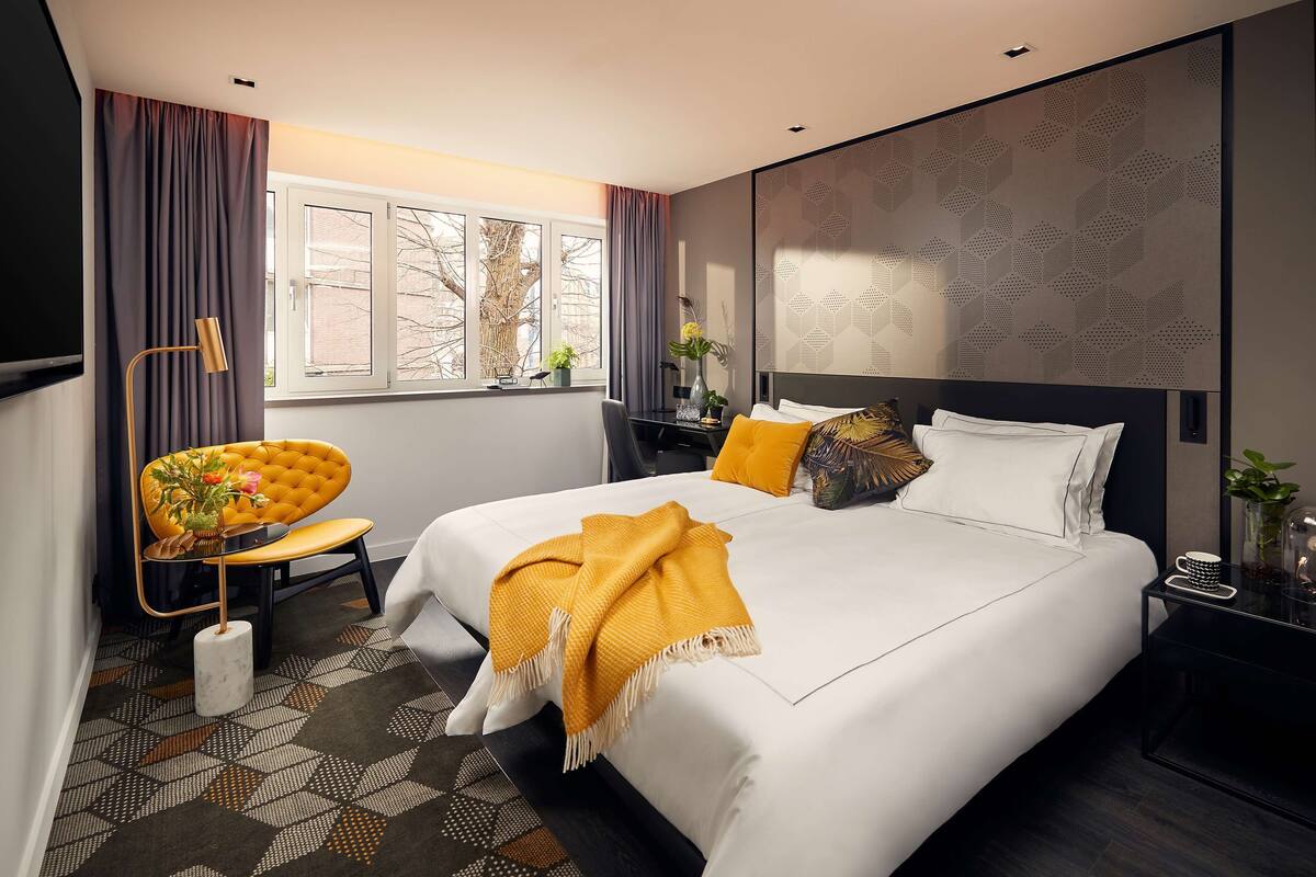Habitación de hotel moderna con una cama grande, un escritorio y una silla. Se ve una ventana con cortinas, una planta en el escritorio y una alfombra estampada en el suelo.