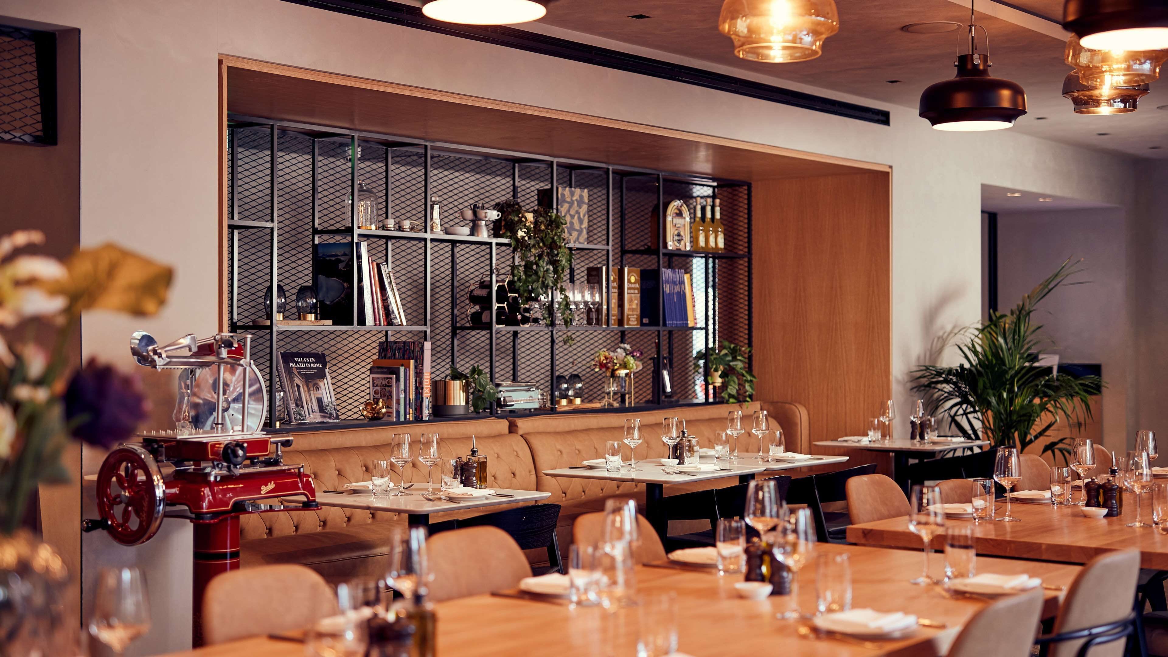 A restaurant with a modern interior, featuring a long counter with multiple tables, a large bookshelf, and a vintage espresso machine.