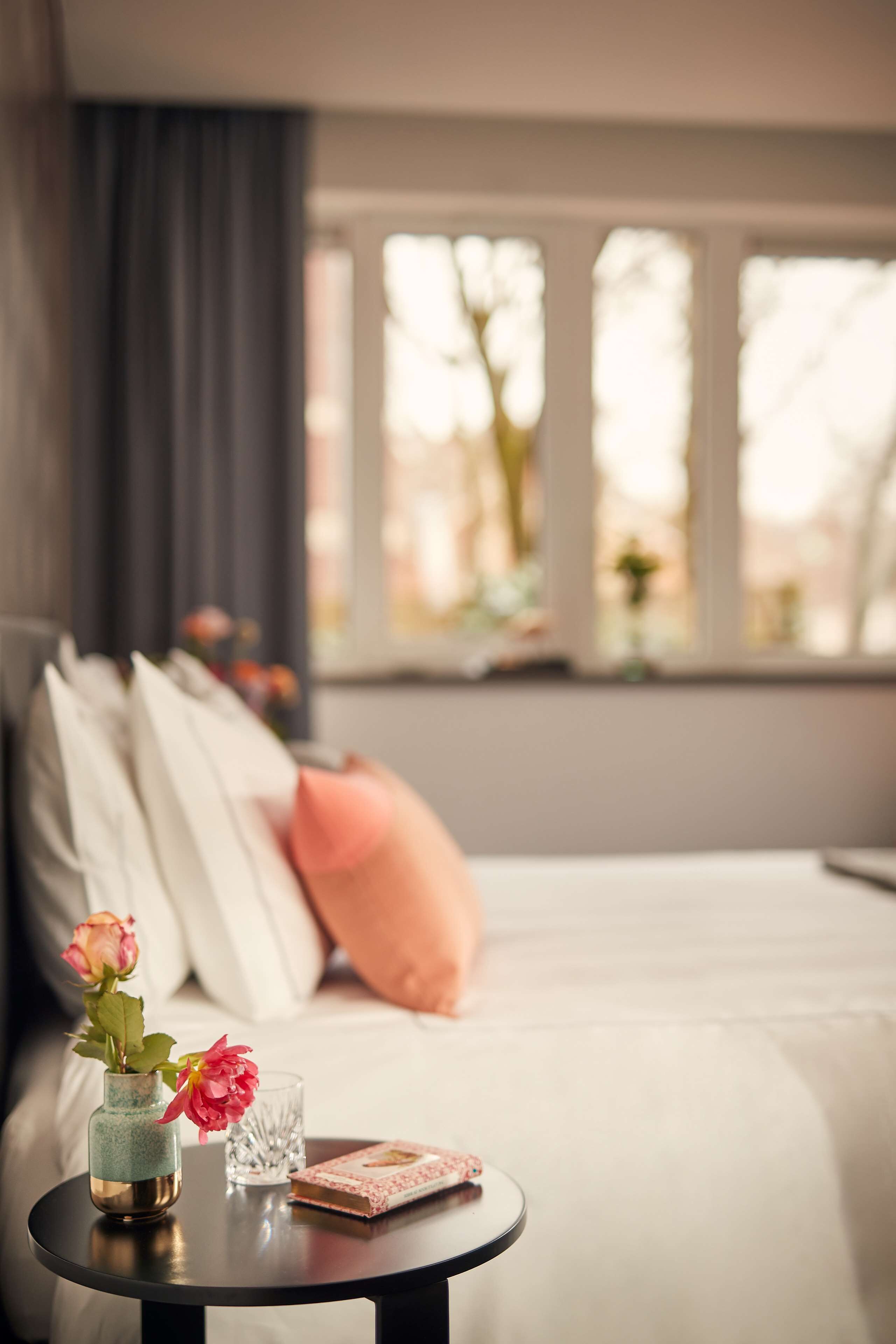 A bedroom with a bed, a bedside table with a vase of flowers, a glass, and a book.