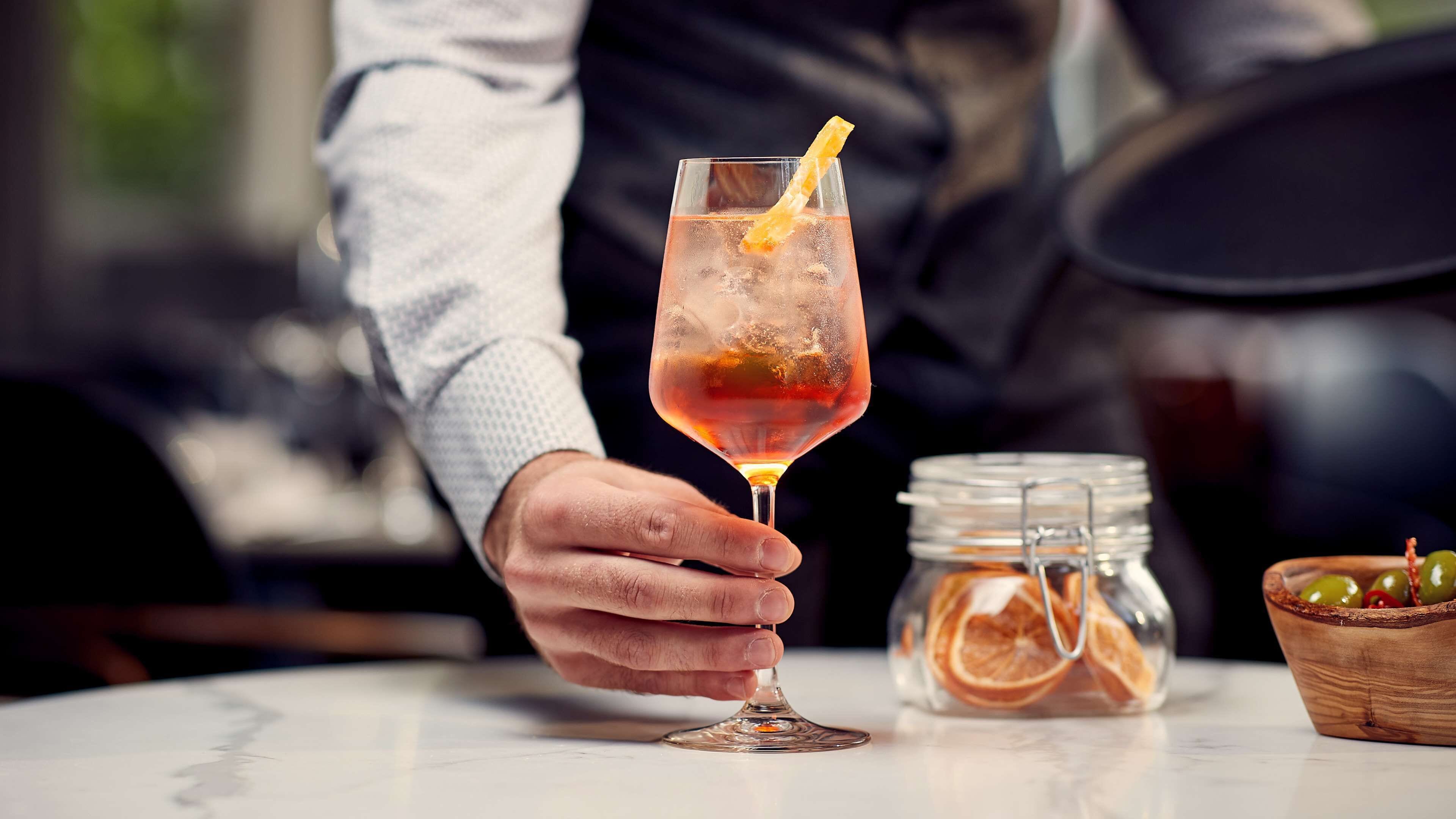 A person holding a cocktail glass with a lemon slice, a jar of dried fruit, and a bowl of olives on a table.