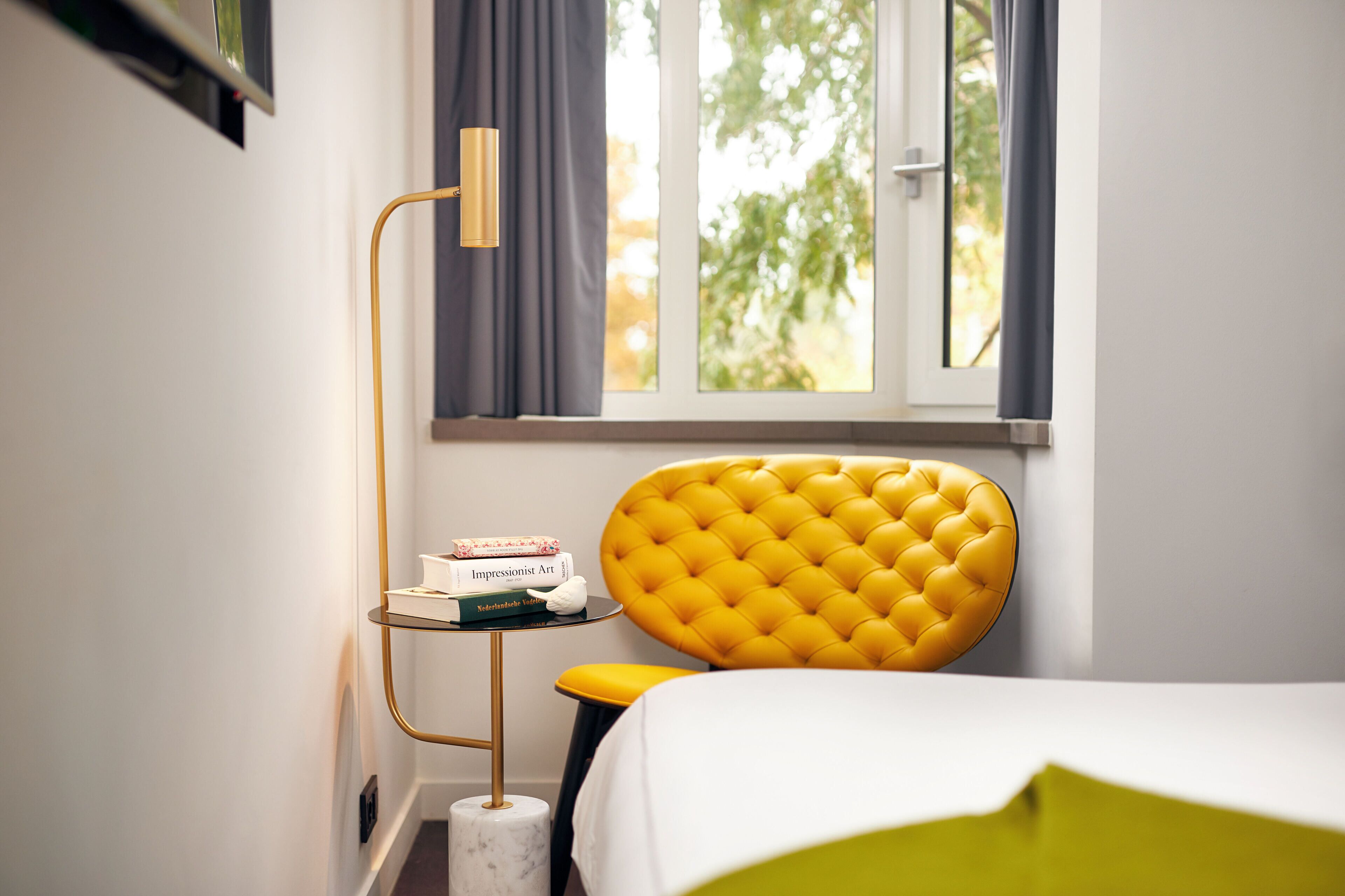 Habitación de hotel con cama, cabecera tapizada en amarillo, mesita de noche con libros, lámpara de pie y ventana con cortinas.