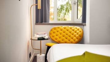 Habitación de hotel con cama, cabecera tapizada en amarillo, mesita de noche con libros, lámpara de pie y ventana con cortinas.
