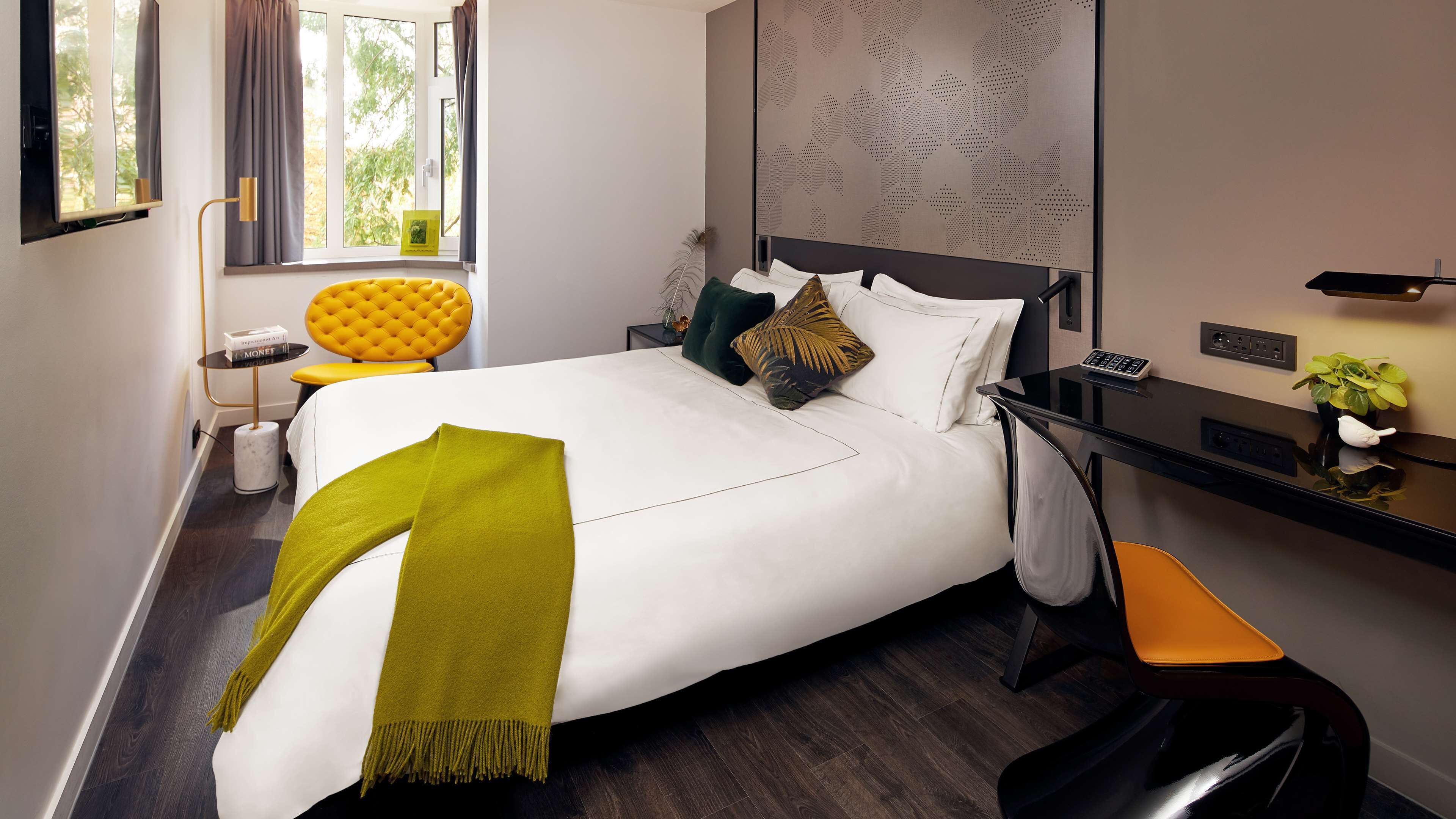 A modern hotel room with a neatly made bed, a desk with a phone, and a view of the outdoors through the window.