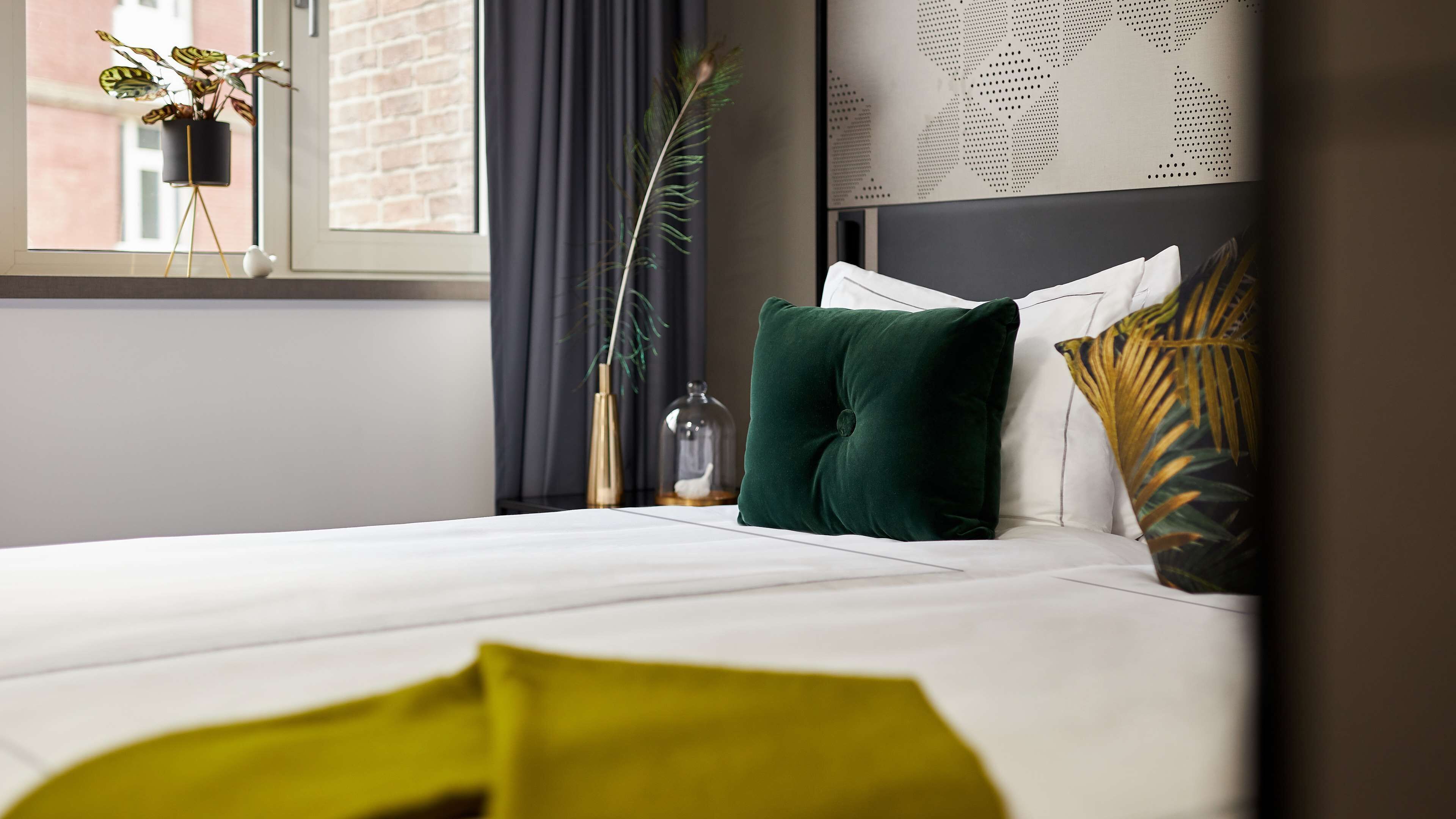 A neatly made bed with white linens, a green pillow, and a decorative plant in a black pot on the windowsill.
