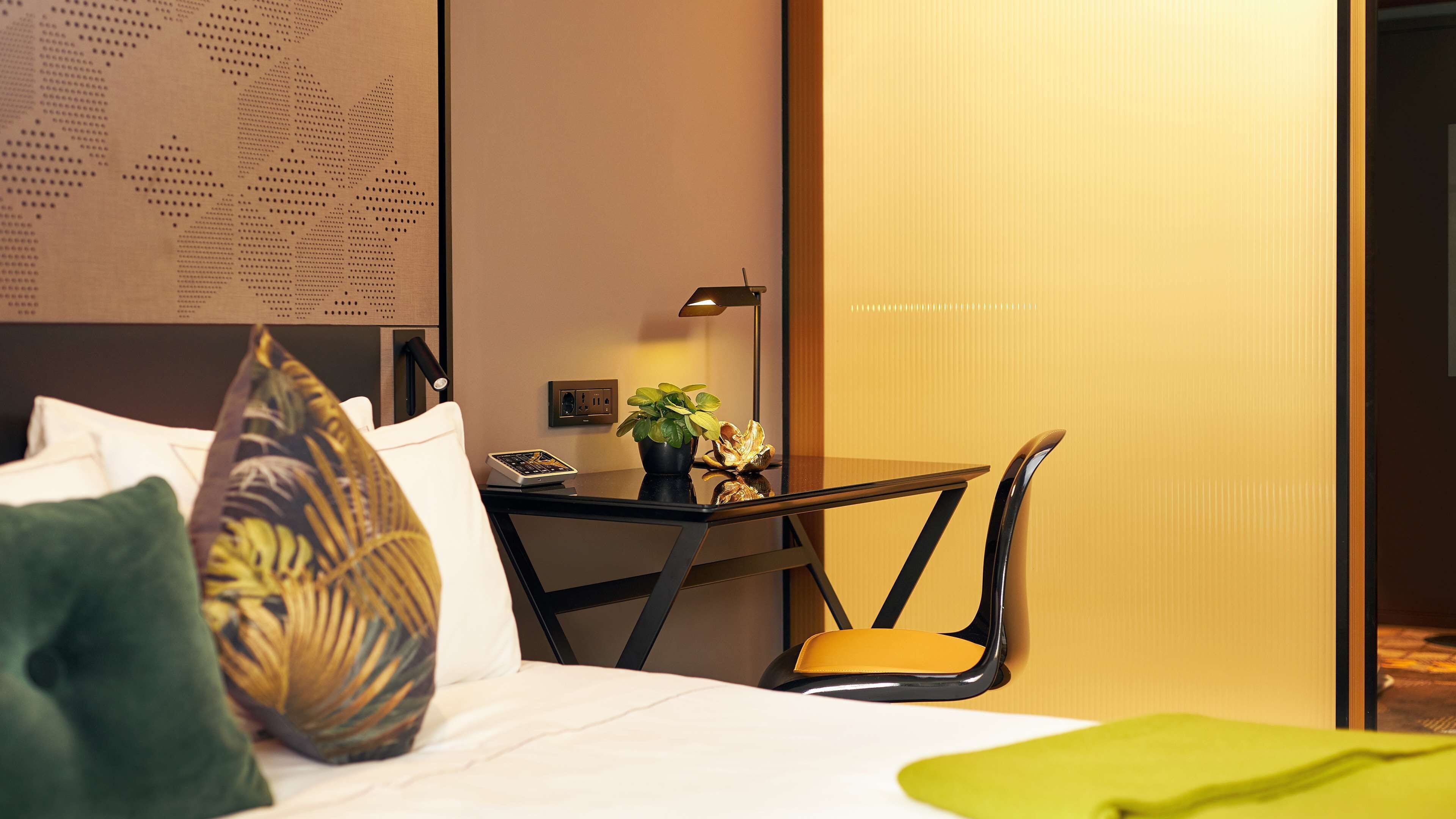 A hotel room with a bed, a desk with a lamp, a chair, and a small table with a plant.