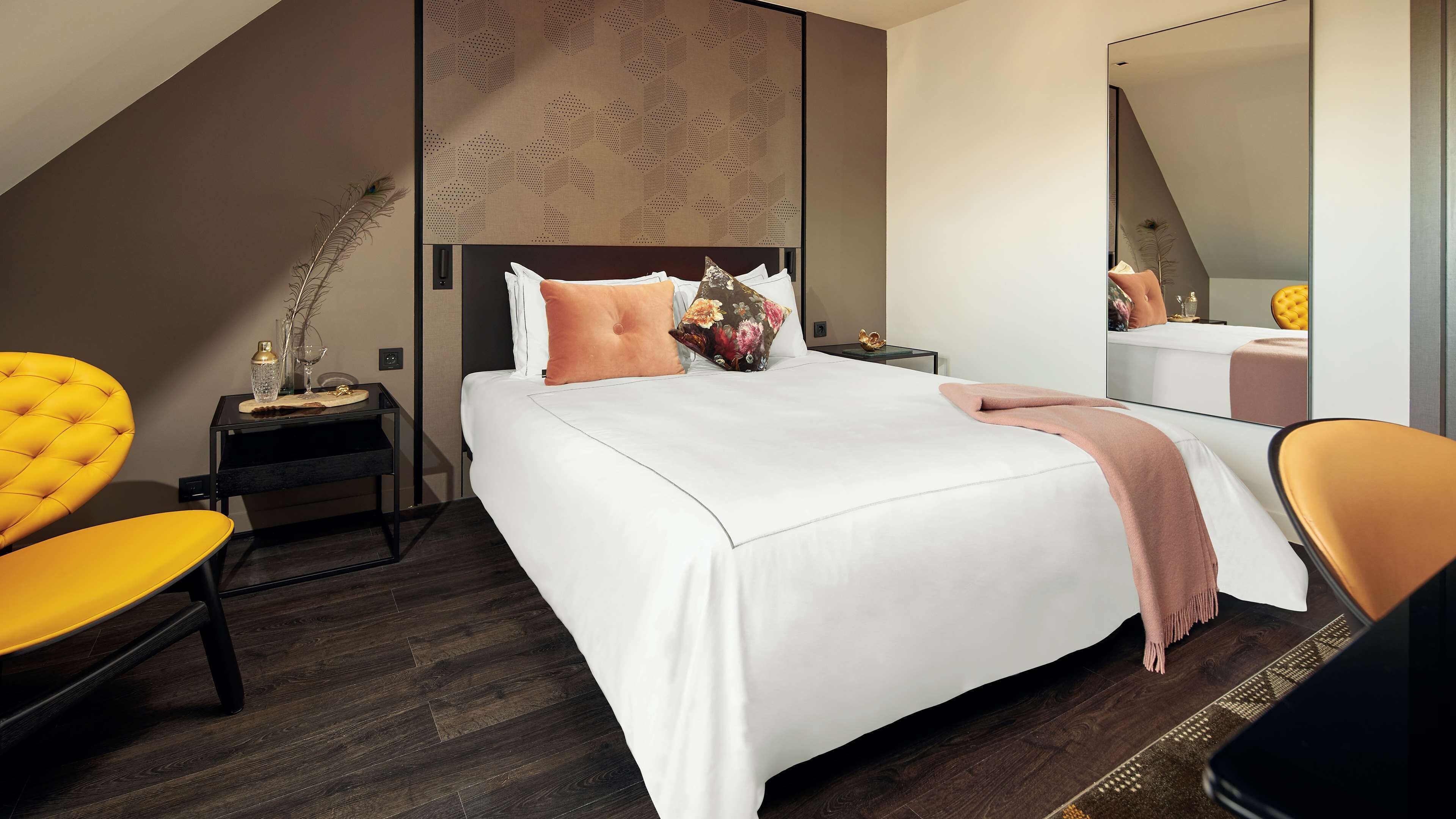 A modern hotel room with a large bed, a side table with a lamp, a mirror, and two chairs.
