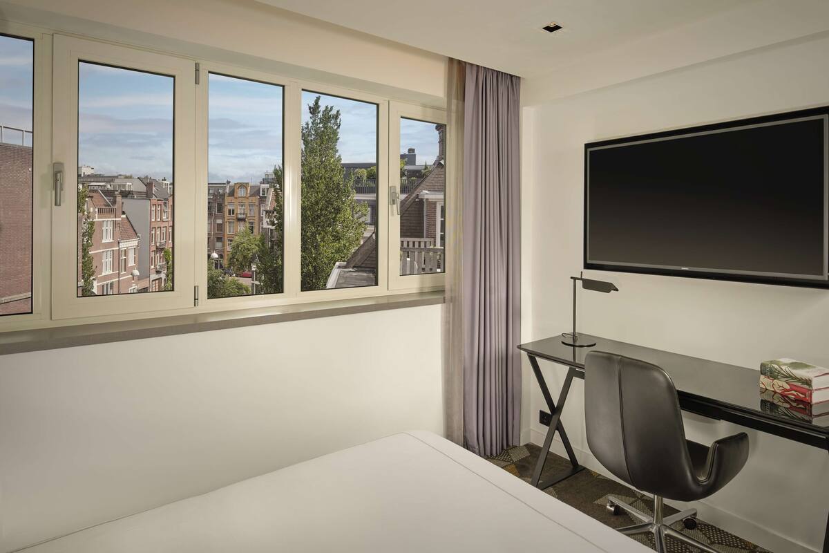 Una habitación de hotel moderna con un ventanal grande, un escritorio con silla, un televisor de pantalla plana y una mesita con libros.