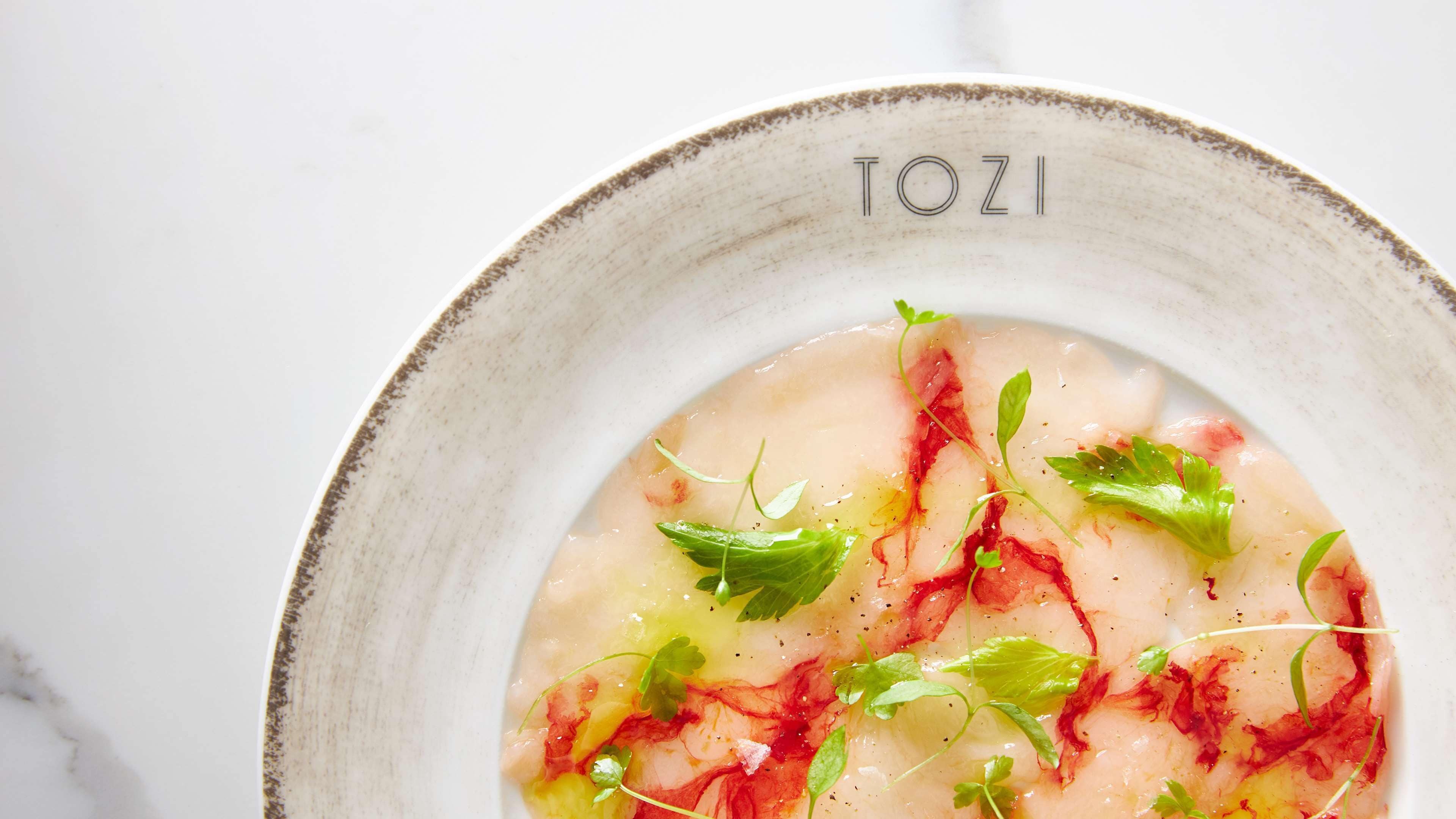Un plato con base cremosa, adornado con hierbas verdes y detalles rojos, servido en un plato de cerámica con la inscripción 'TOZI'.