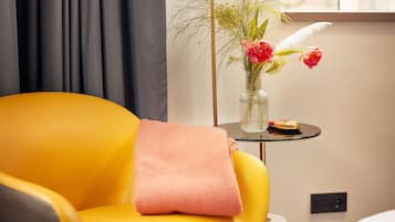 Una poltrona gialla con una coperta rosa, un tavolino rotondo con un vaso di fiori e pasticcini, e una finestra con tende.