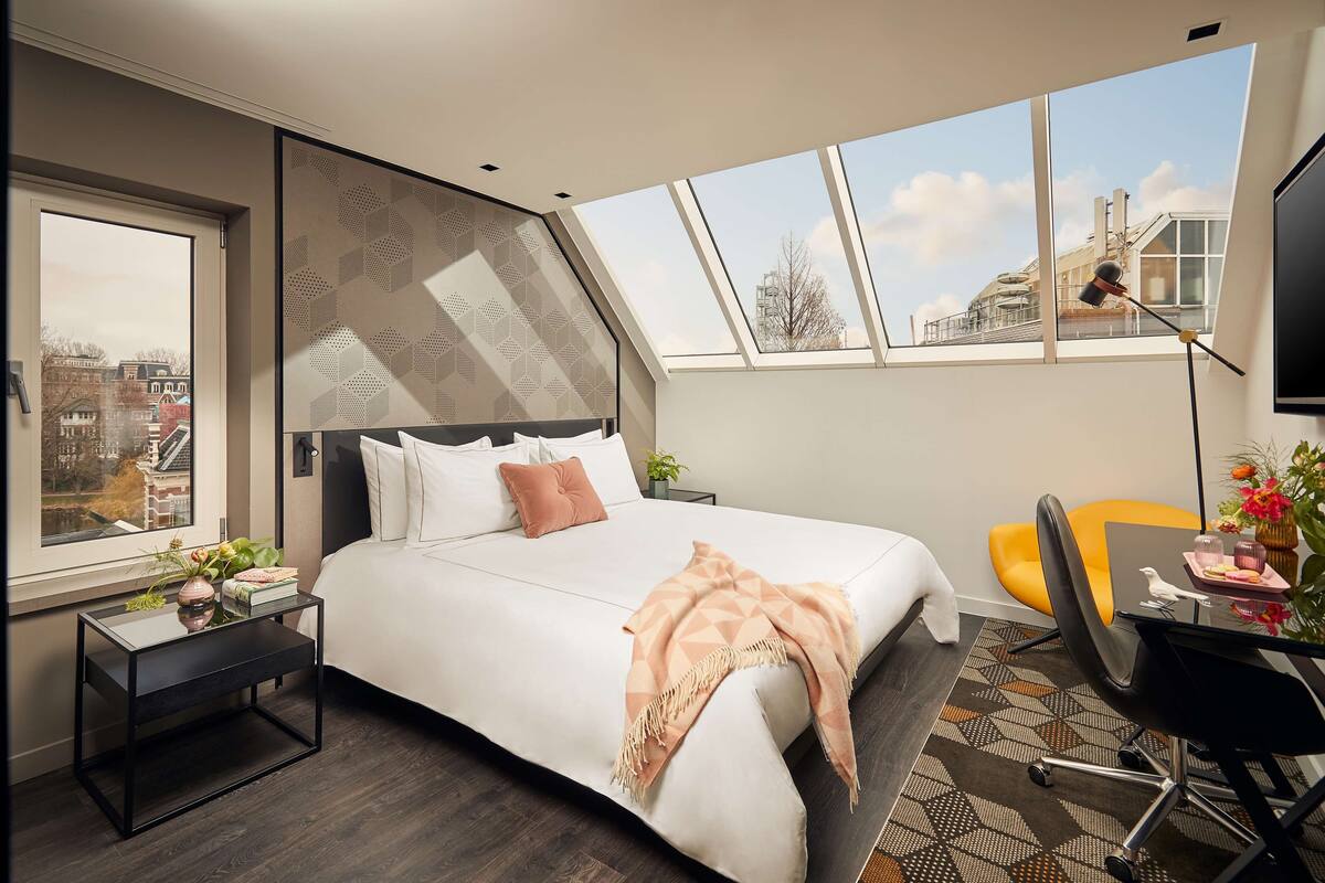 Un dormitorio moderno con una cama grande, un escritorio y una silla. Hay una ventana con vistas a edificios.