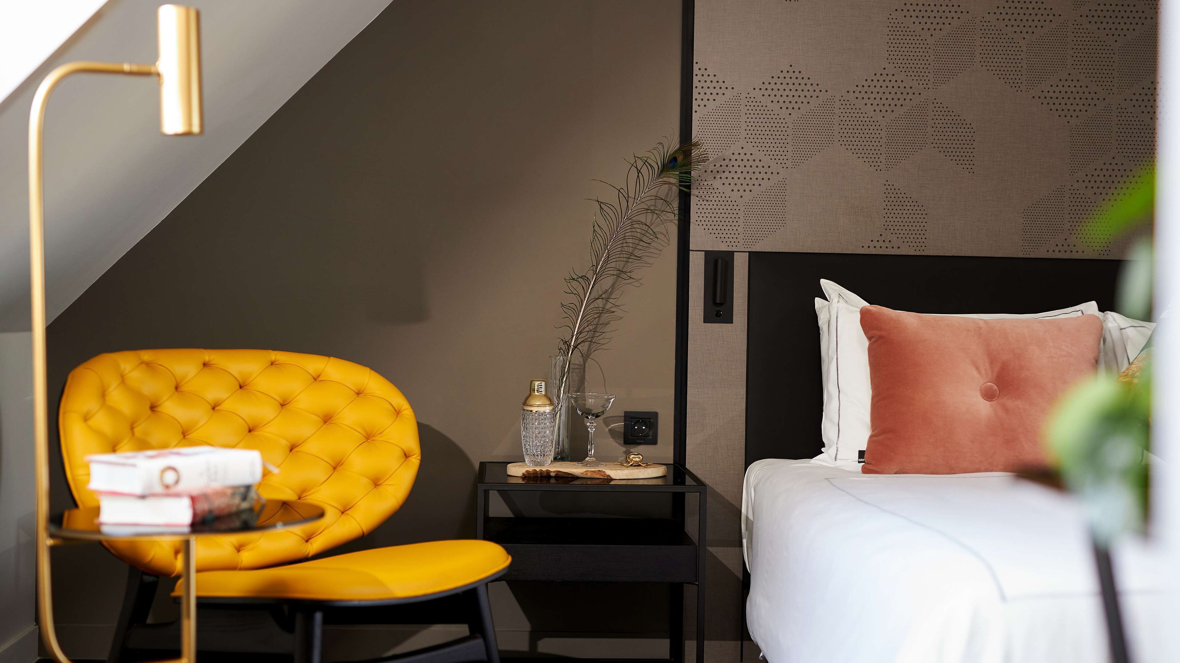 Habitación de hotel con una butaca amarilla acolchada, una mesita de noche negra con un jarrón y una botella, una cama con almohadas blancas y rosas, y un papel tapiz estampado.