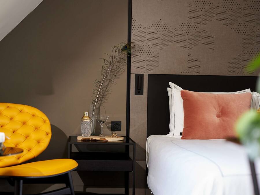 Habitación de hotel con una butaca amarilla acolchada, una mesita de noche negra con un jarrón y una botella, una cama con almohadas blancas y rosas, y un papel tapiz estampado.