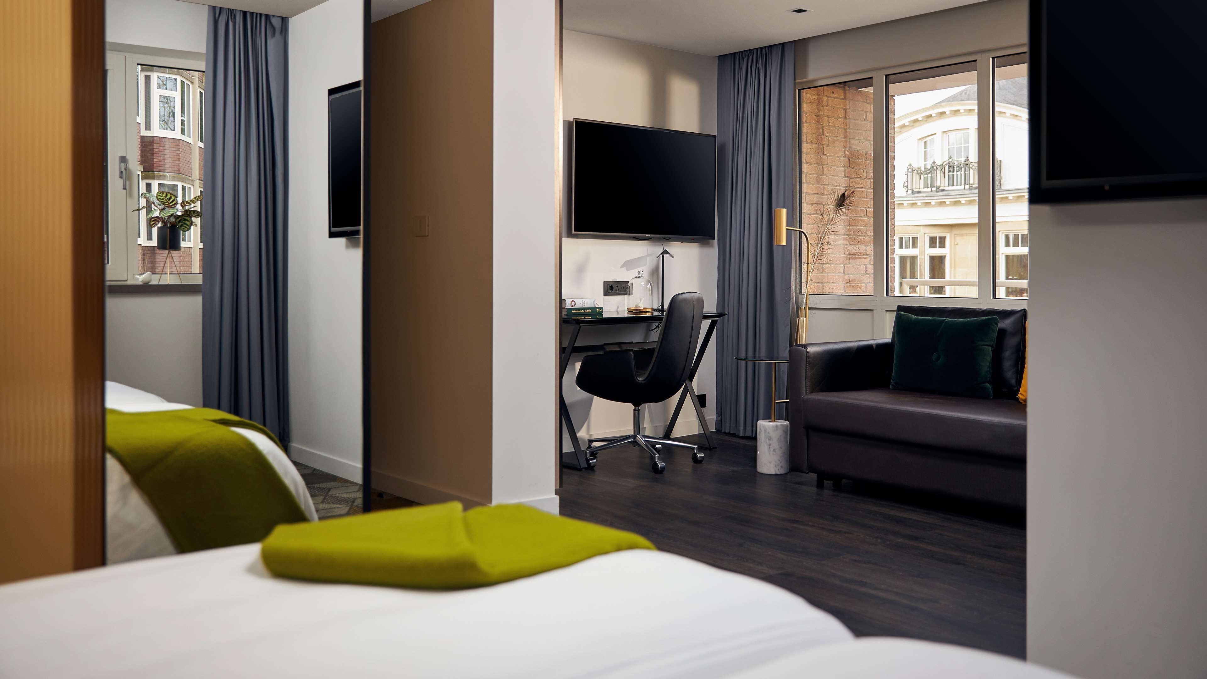 Una habitación de hotel con una cama, un escritorio con silla, un sofá y un televisor montado en la pared.