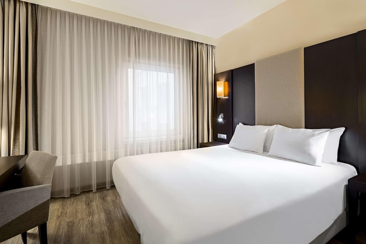 A hotel room with a large bed, a desk, a chair, a nightstand, and a window with curtains.