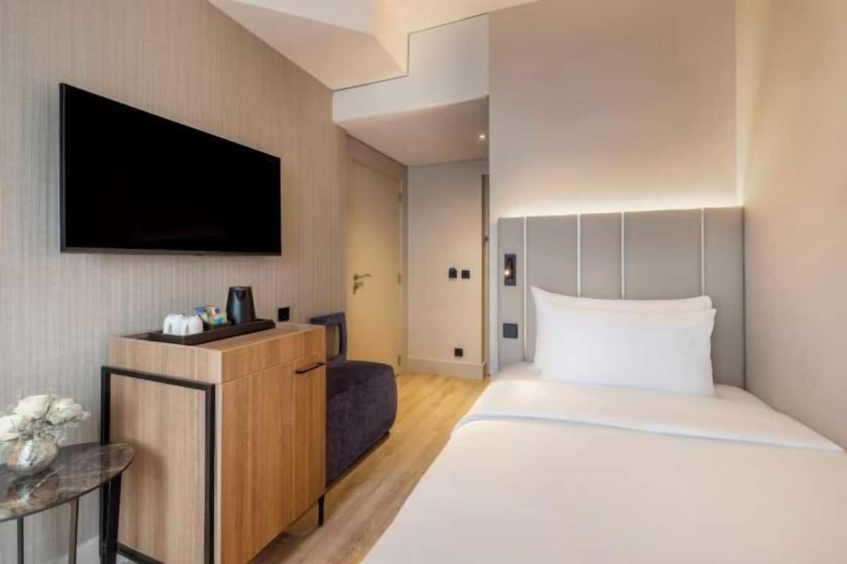 A modern hotel room with a flat-screen TV, a wooden desk with a chair, a small table with a vase, and a bed with a headboard.