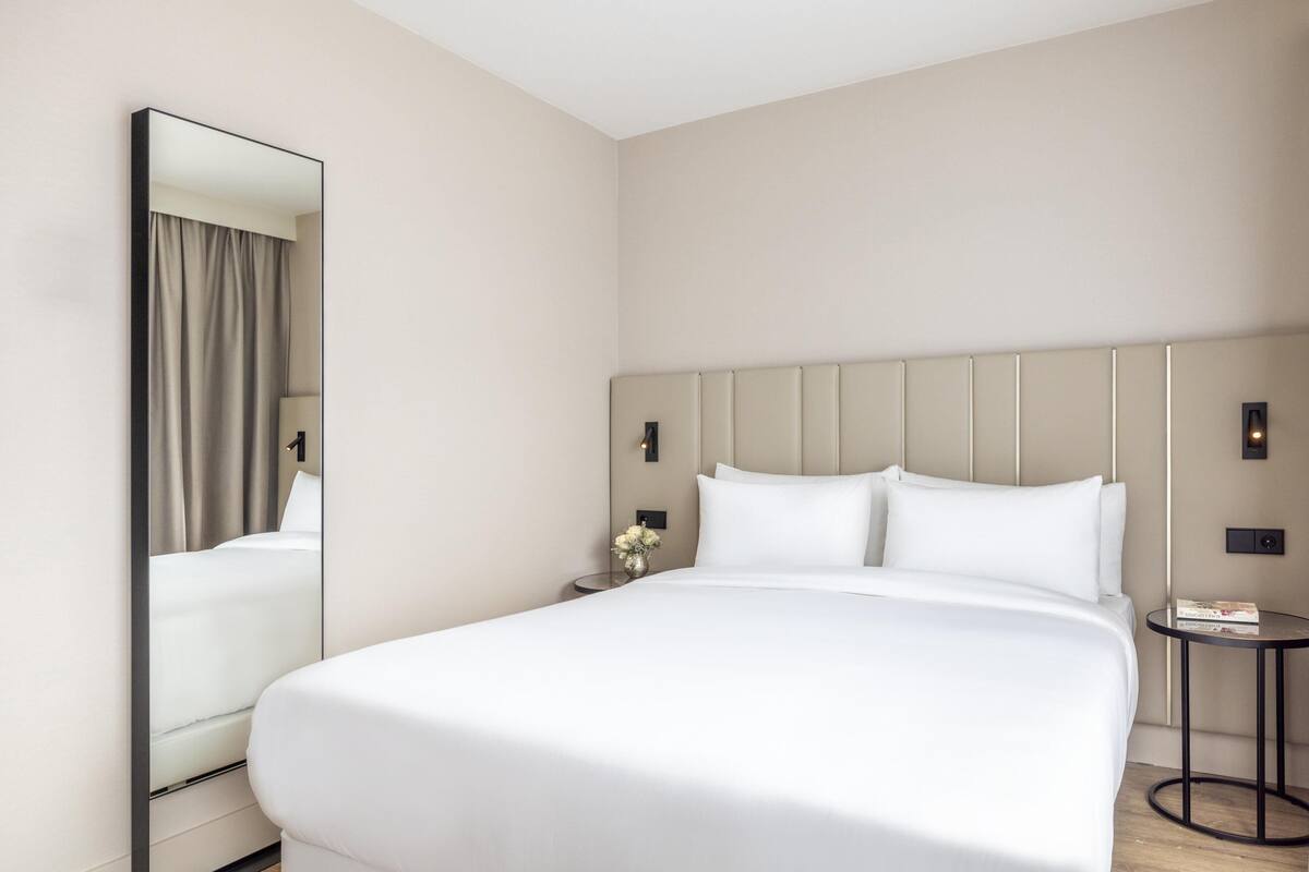A modern hotel room with a large bed, a bedside table with a vase of flowers, a round side table, and a large mirror.