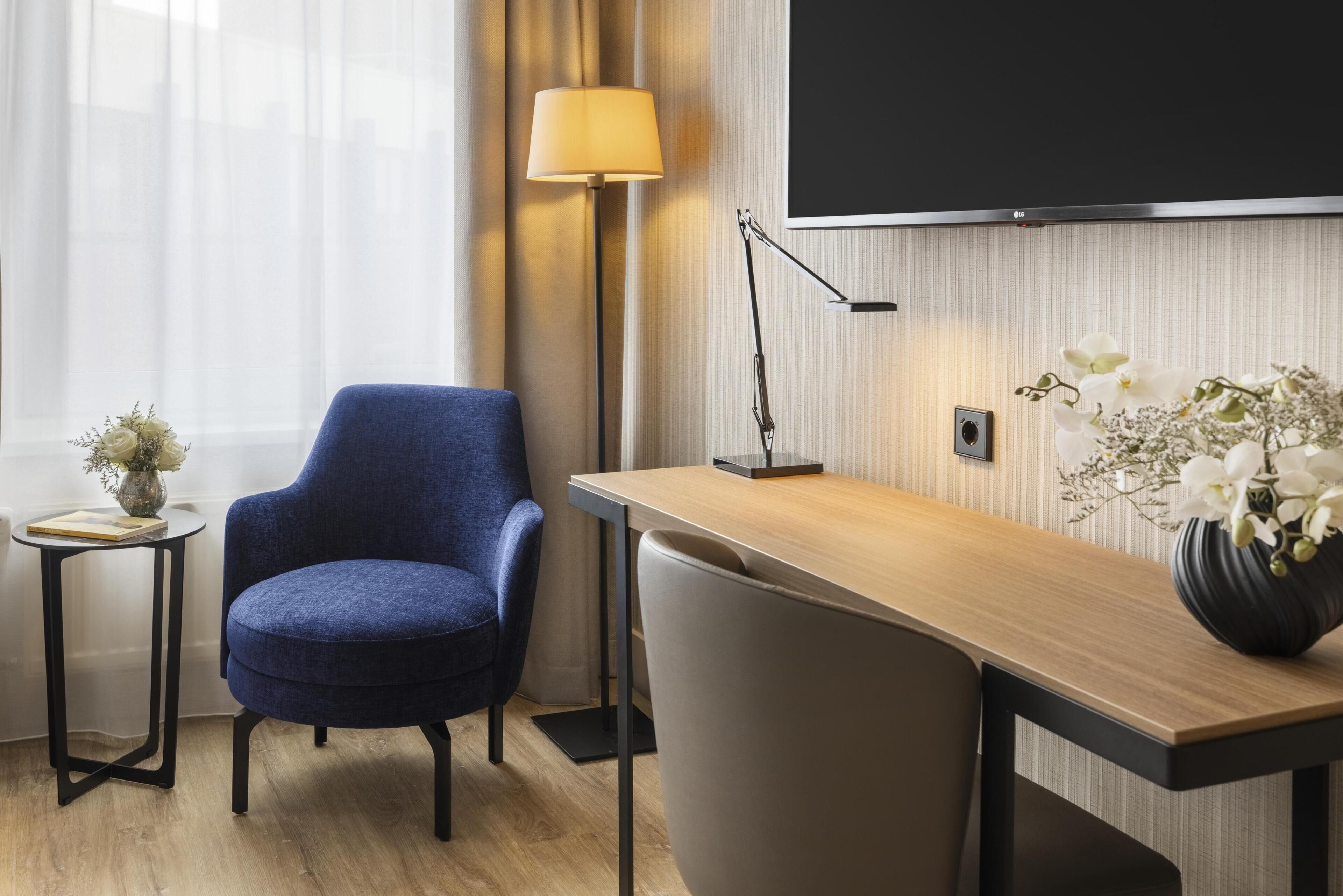 A modern hotel room with a blue armchair, a wooden desk, a lamp, a TV, and a vase with flowers.