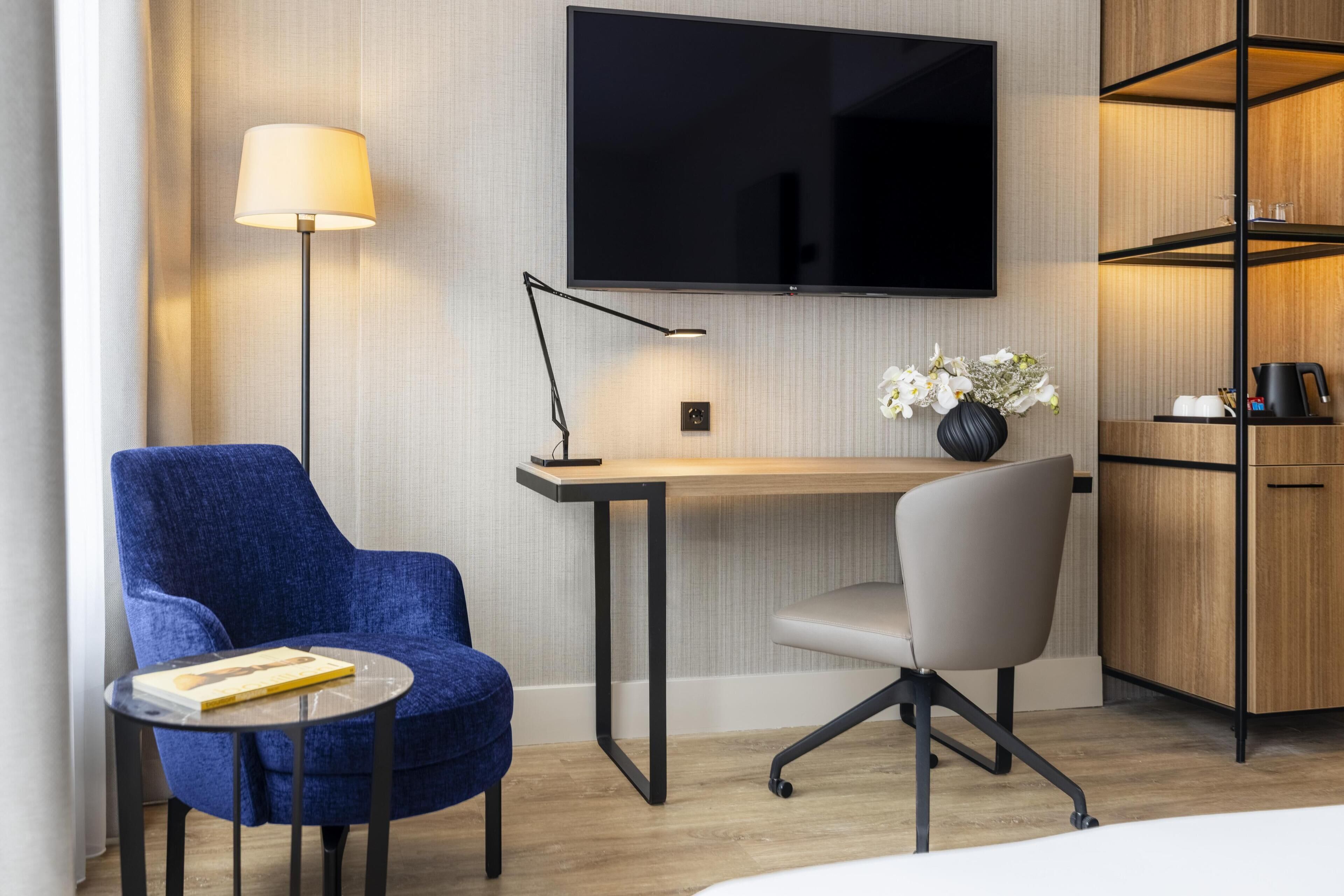 A modern hotel room with a flat-screen TV, a desk with a lamp, a blue armchair, a grey office chair, and a wooden cabinet.