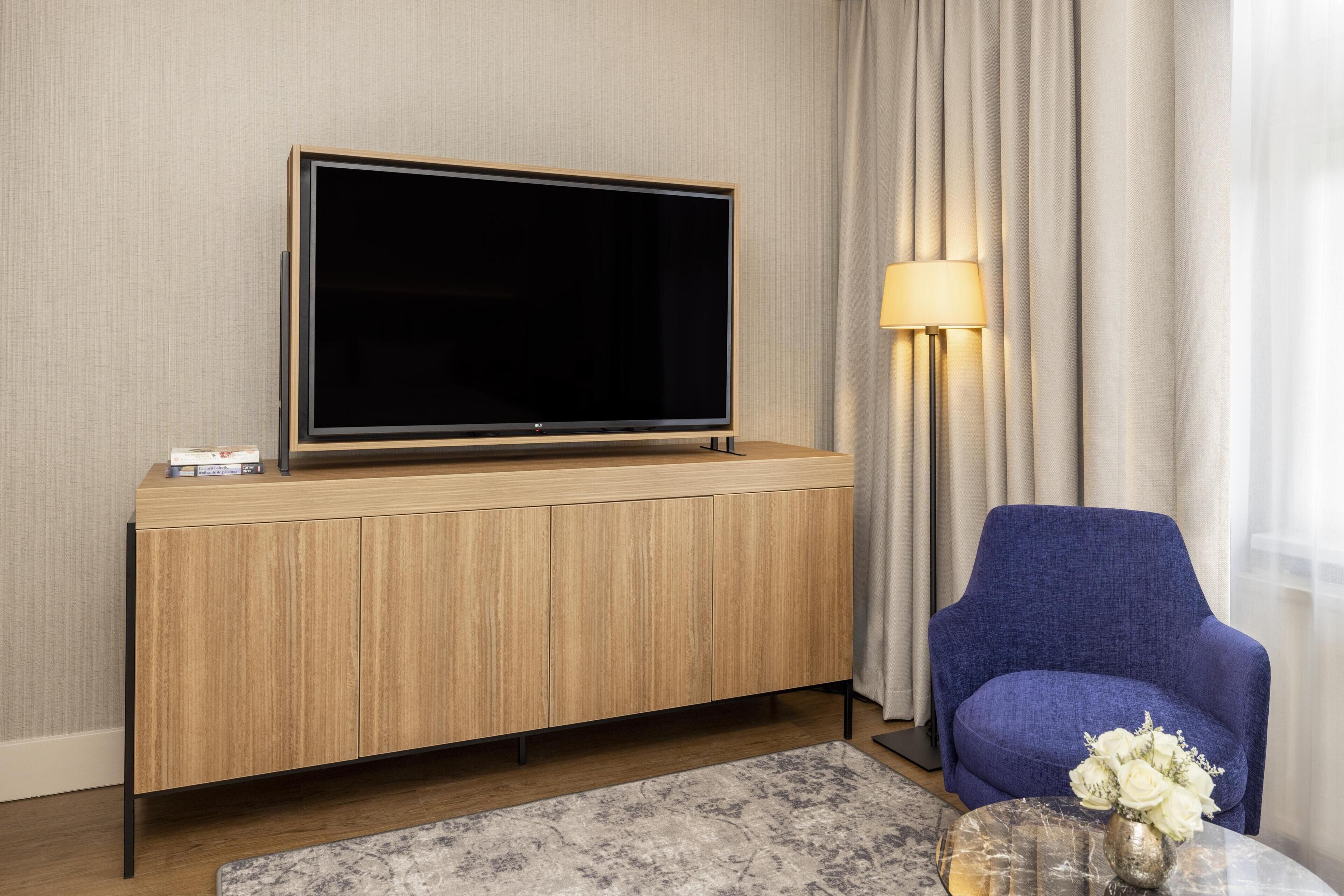 Ein modernes Wohnzimmer mit einem Flachbildfernseher auf einer Holzkommode, einem blauen Sessel, einer Lampe und einer Vase mit Blumen auf einem Glastisch.