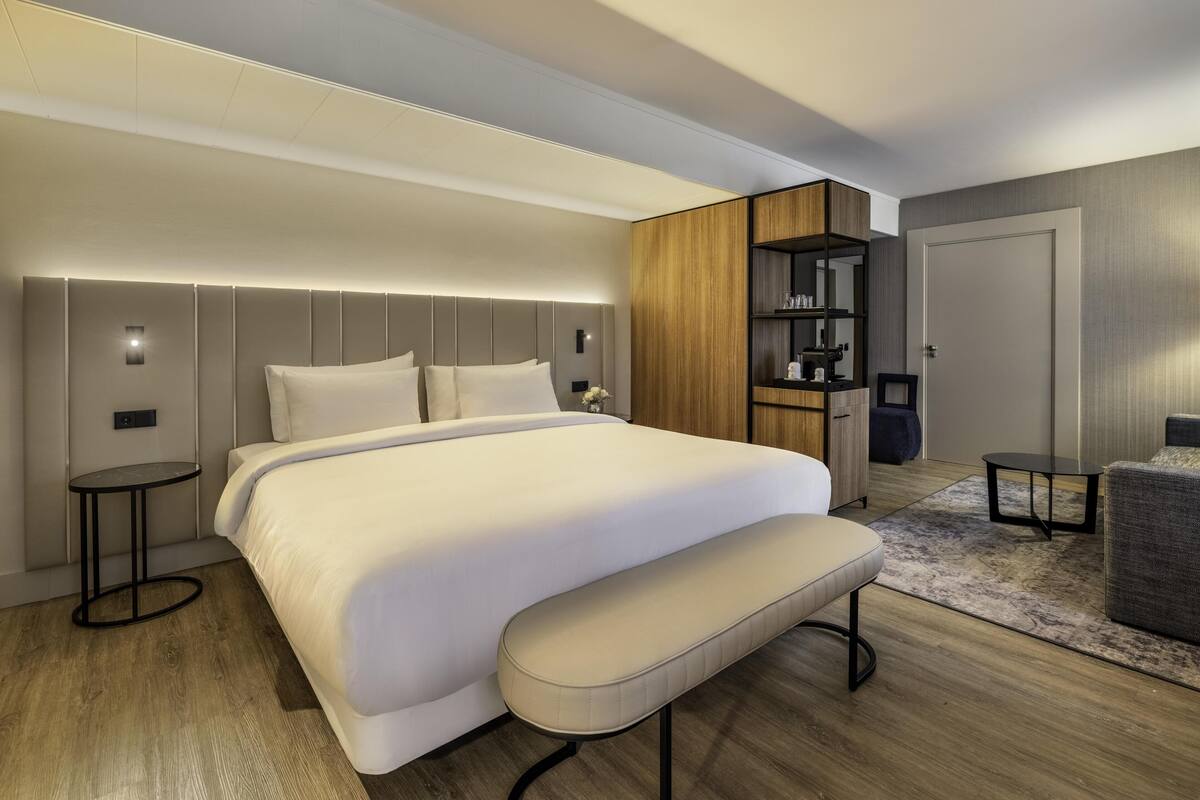 A modern bedroom with a large bed, a bench, a bedside table, a built-in wardrobe, and a small seating area.