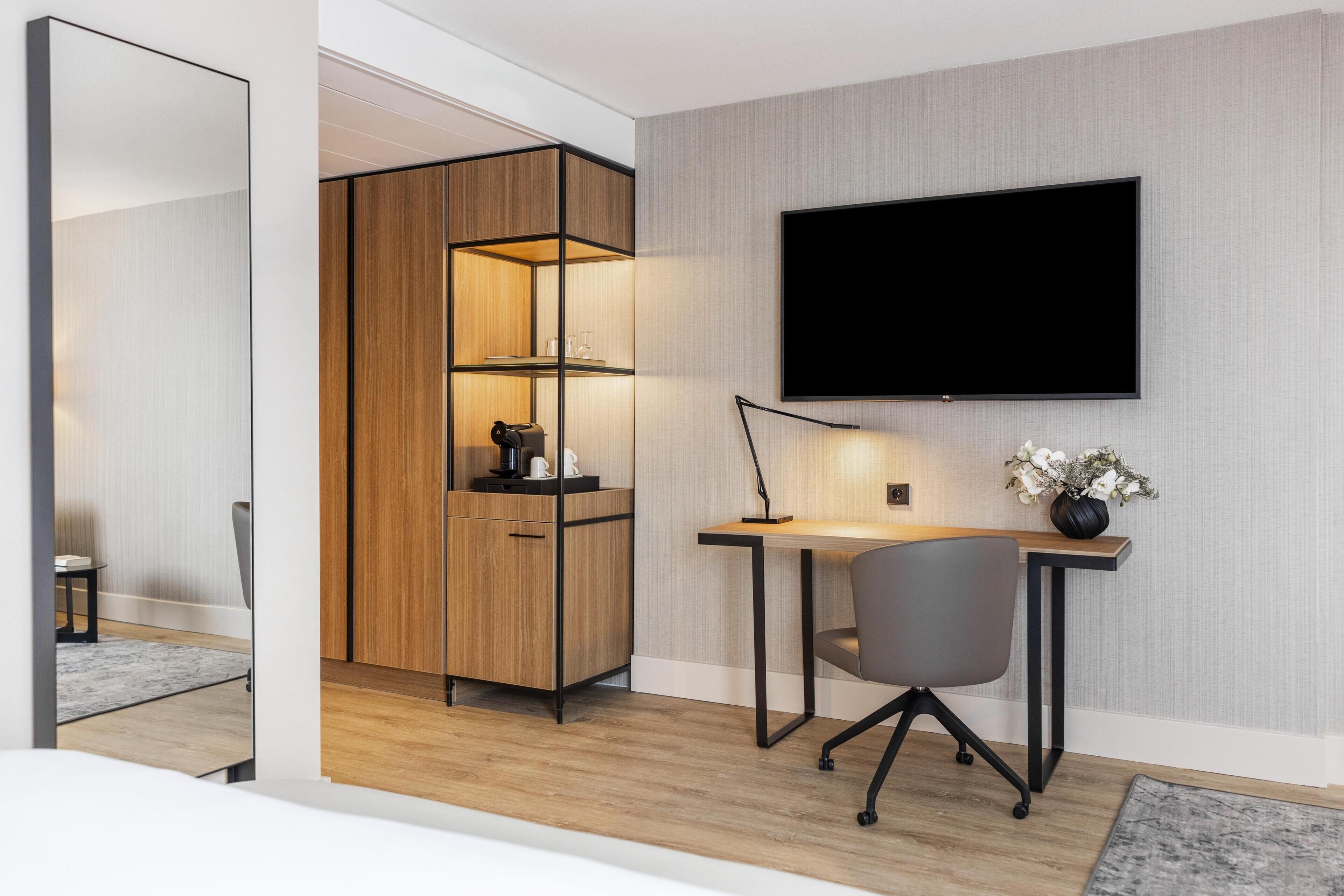 A modern bedroom with a wooden wardrobe, a desk with a chair, a flat-screen TV, and a bedside table with a vase of flowers.