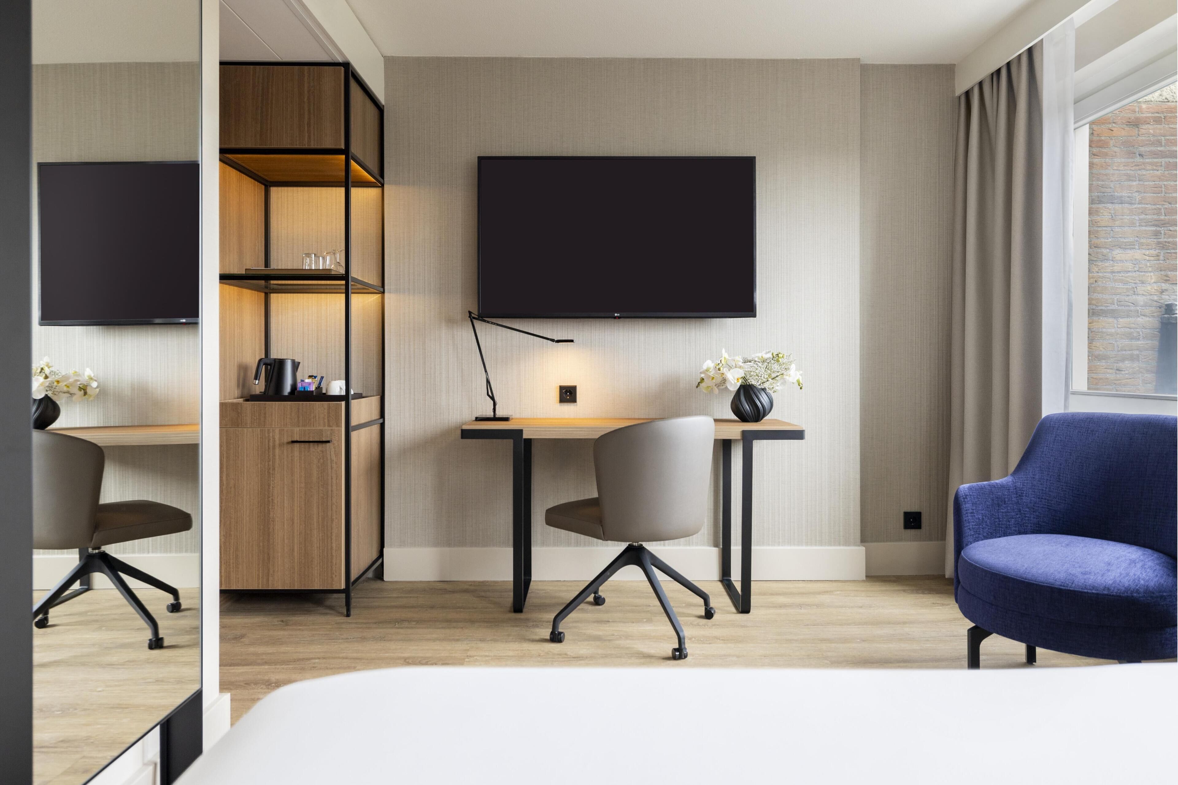 A modern hotel room with a bed, a desk, a chair, a TV, and a wardrobe.