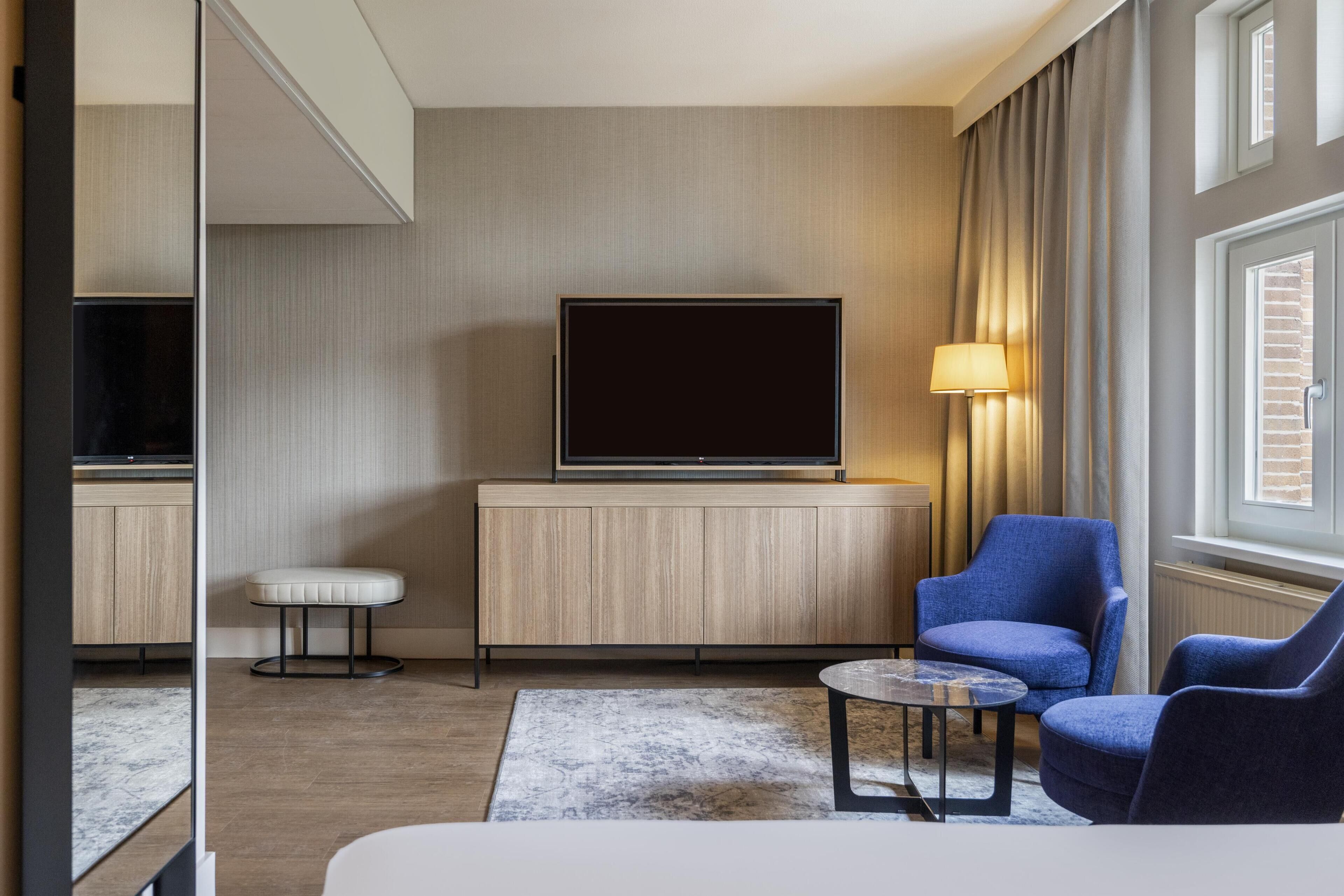 A modern hotel room with a flat-screen TV on a wooden cabinet, a blue armchair, a small round table, and a large window with curtains.