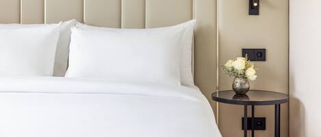Een opgemaakt bed met witte lakens en een vaas bloemen op een klein, rond tafeltje ernaast.
