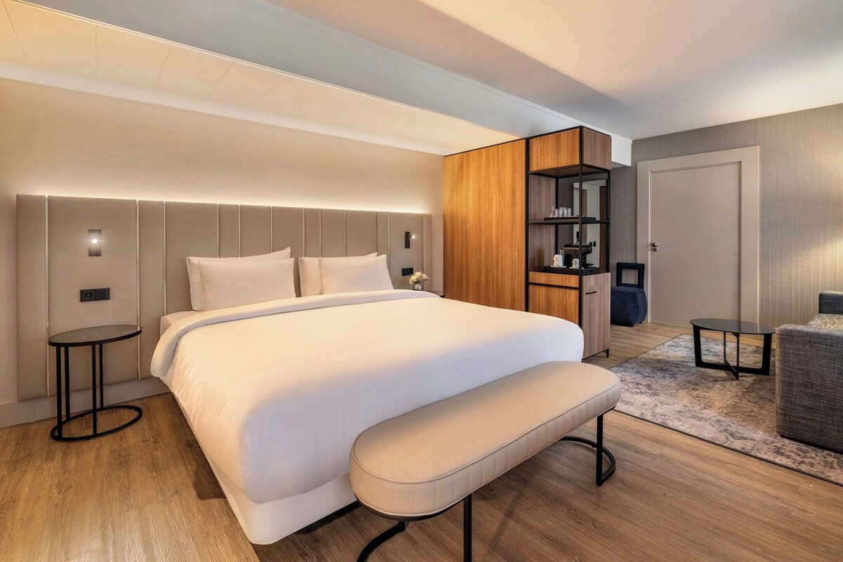 Una habitación de hotel moderna con una cama grande, mesitas de noche, una banca, armario empotrado y una zona de estar.