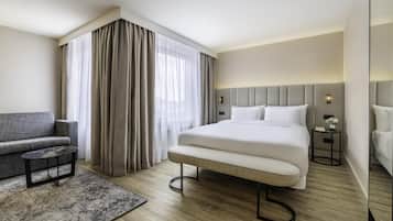 Una habitación de hotel moderna con una cama grande, un sofá gris, una mesita auxiliar pequeña y un ventanal con cortinas.