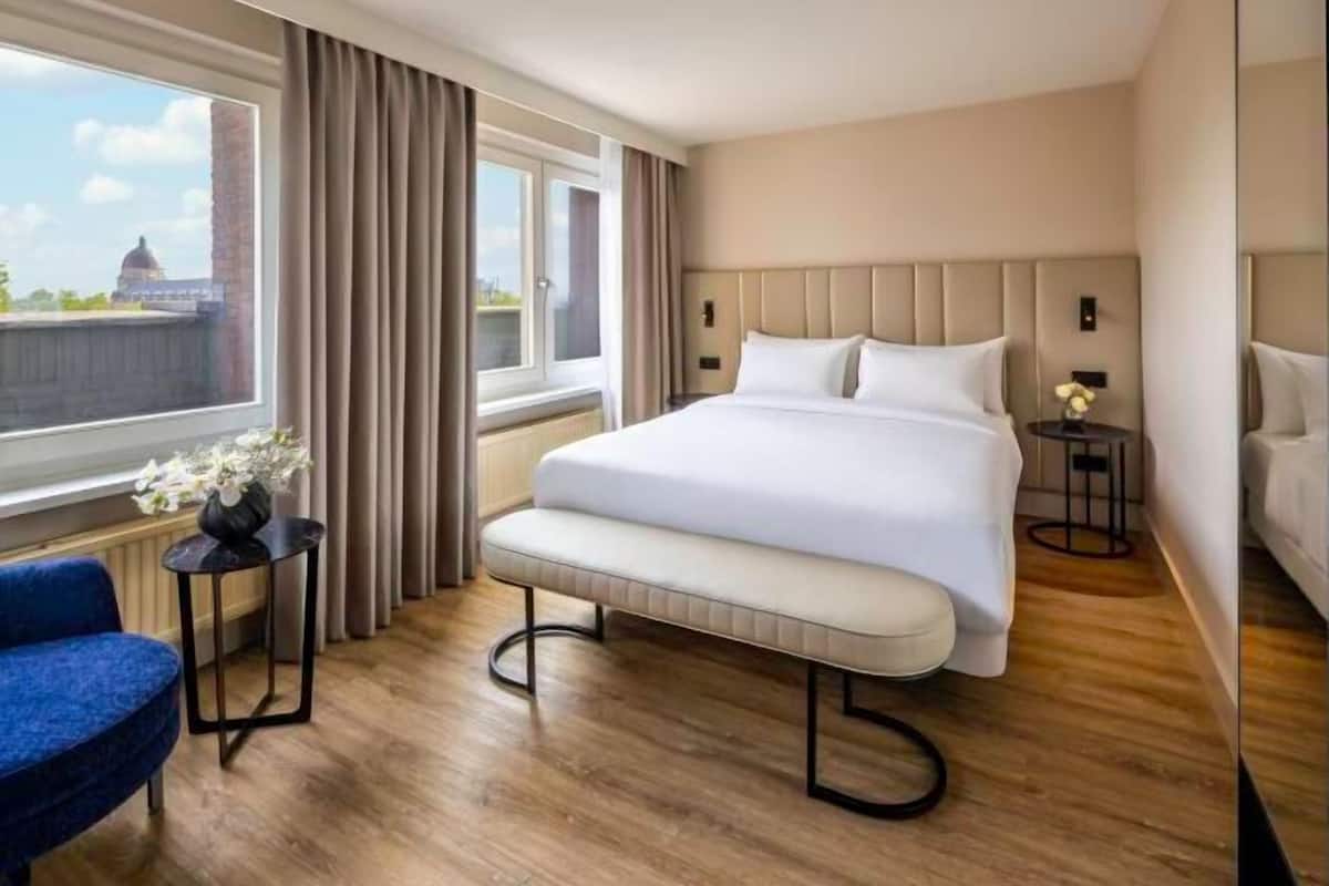 A hotel room with a large bed, a bench, a small table with a vase, a blue chair, and a view of a building through the window.