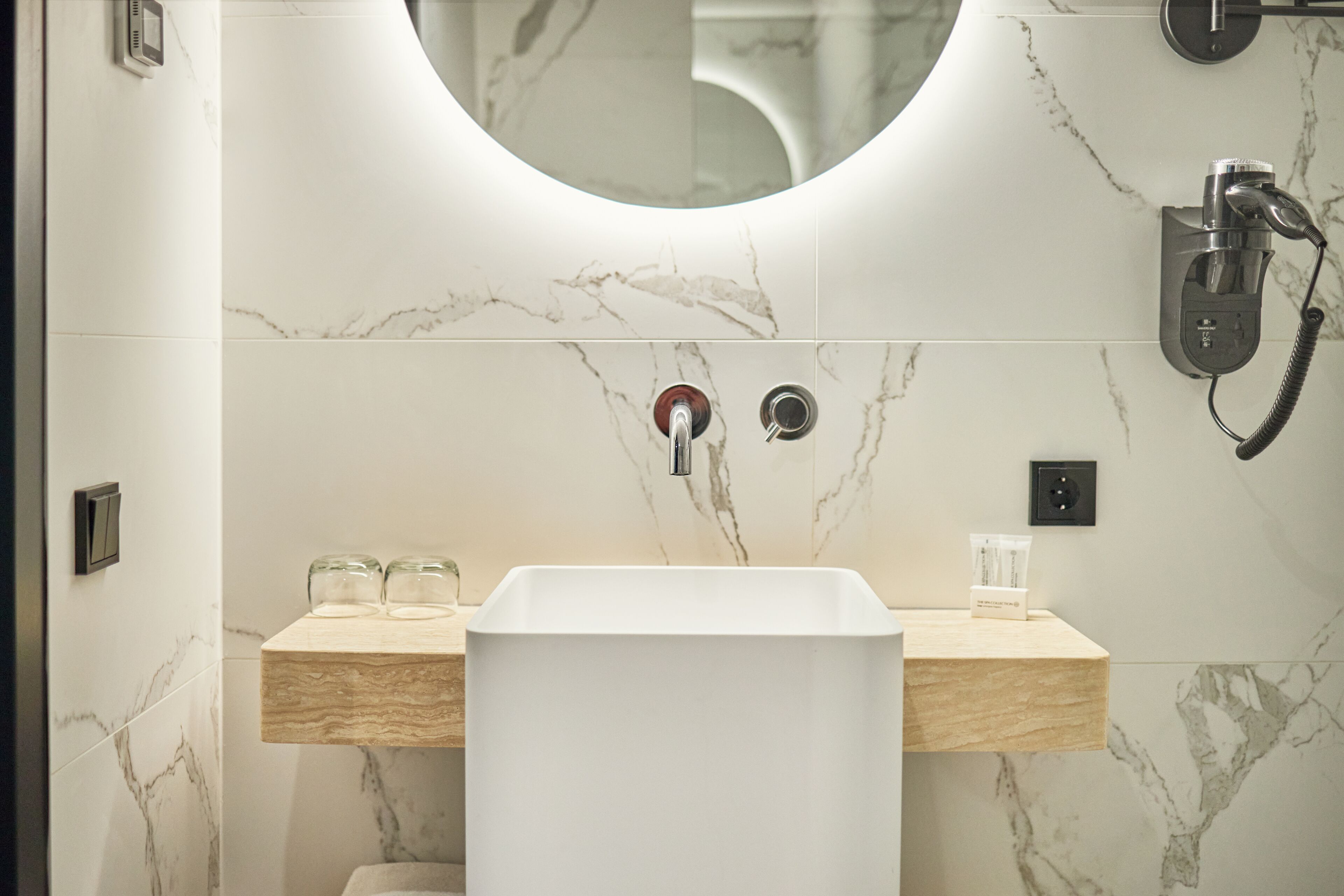 Un baño con lavamanos blanco, paredes de mármol y un espejo redondo.