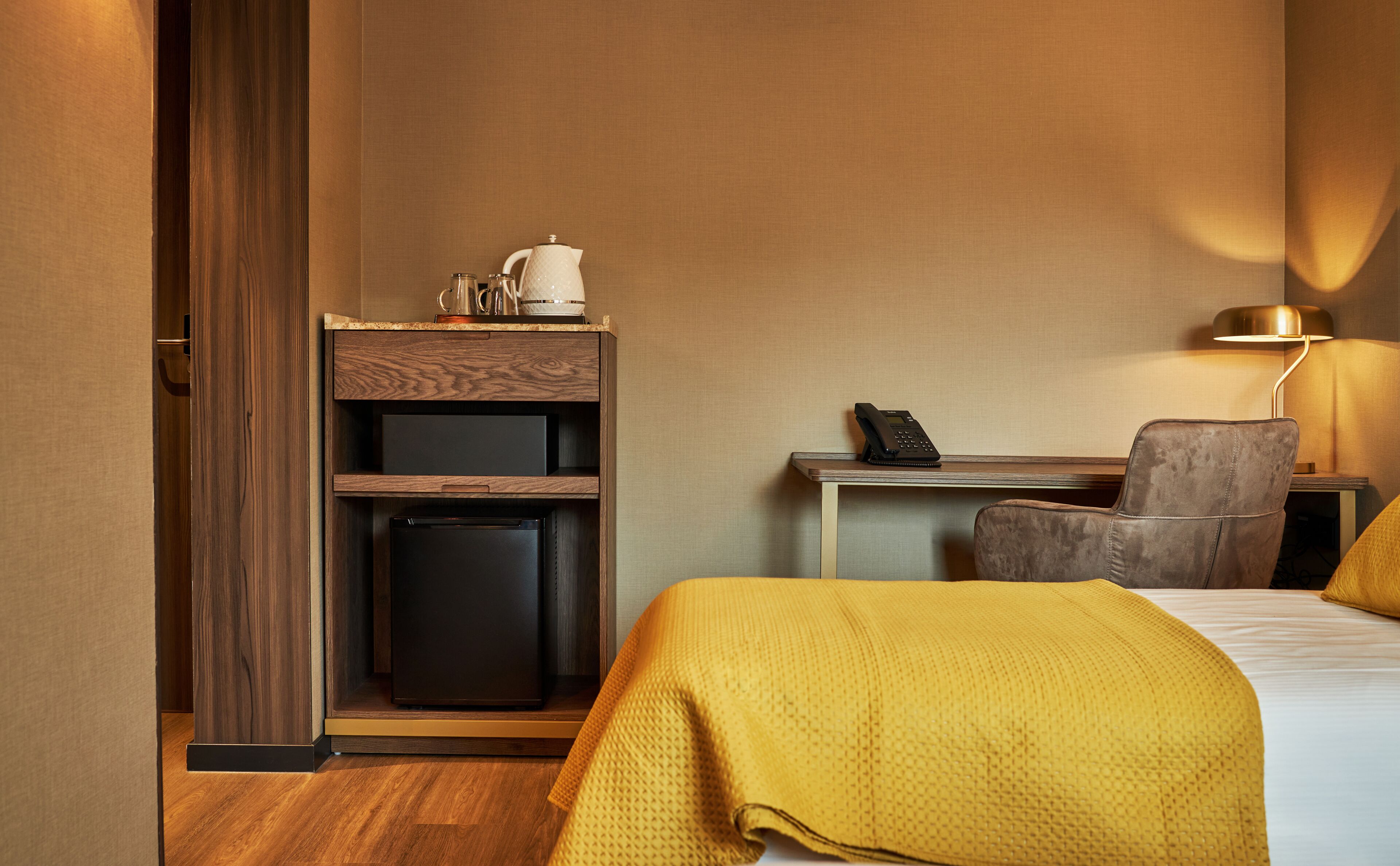 Ett hotellrum med en säng, ett skrivbord med en telefon, en stol, ett litet kylskåp och en kaffebryggare.