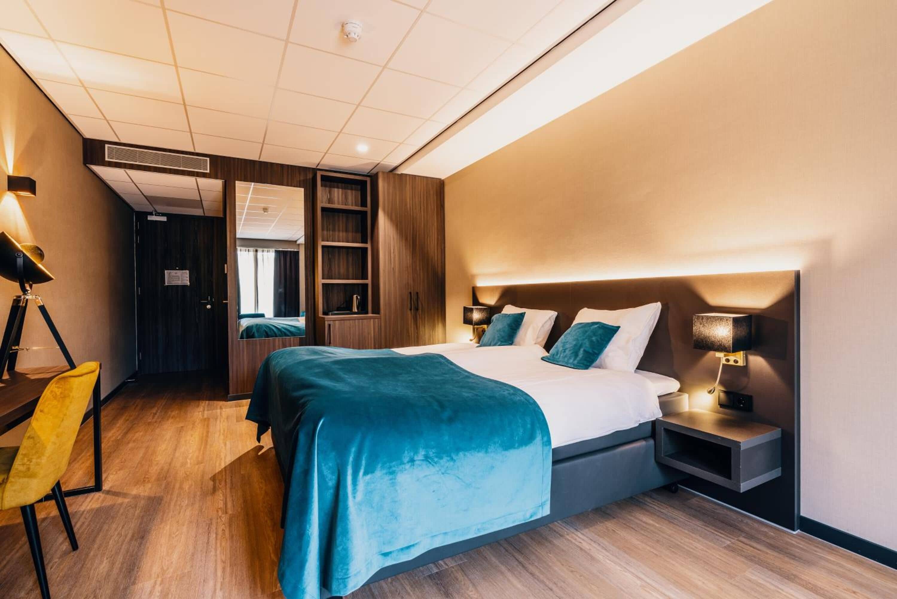 Ett modernt hotellrum med en stor säng, ett skrivbord och en stol. Det finns en garderob och ett litet bord med en lampa.