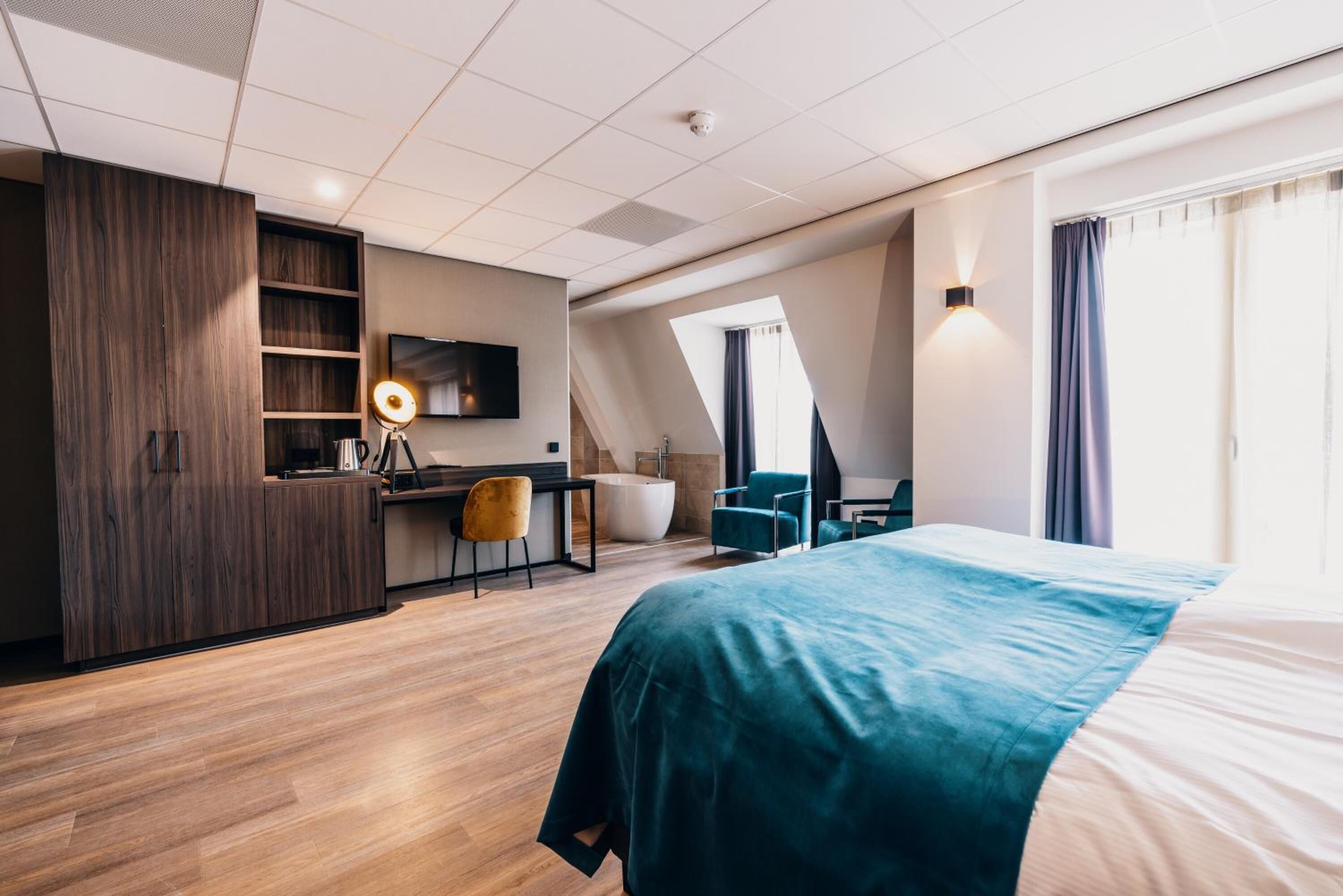 A modern hotel room with a large bed, a desk with a chair, a vanity with a mirror, and a bathtub.