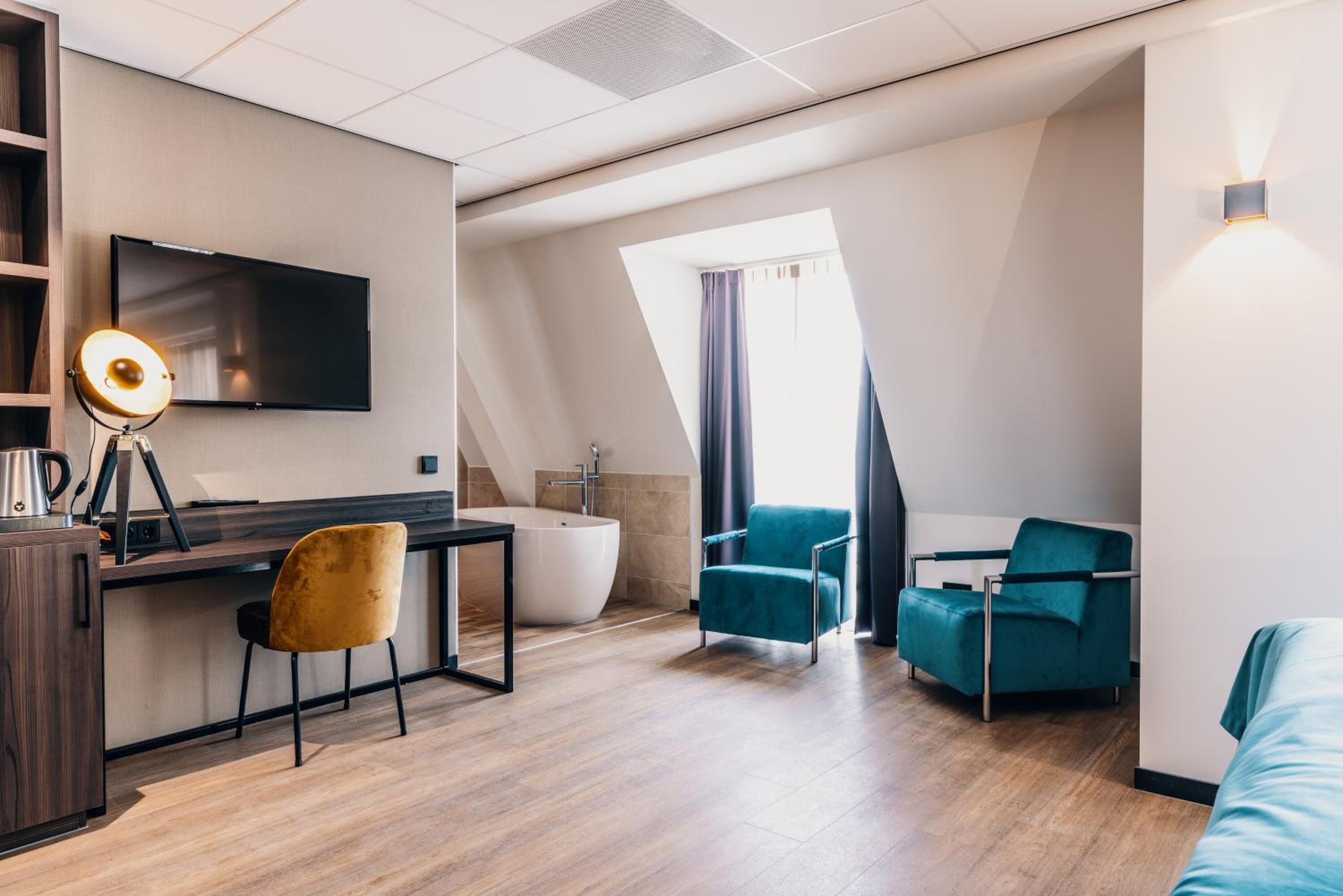 A modern hotel room with a flat-screen TV, a desk with a lamp, a bathtub, and two teal armchairs.