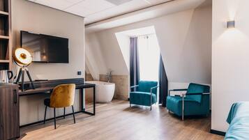 A modern hotel room with a flat-screen TV, a desk with a lamp, a bathtub, and two teal armchairs.