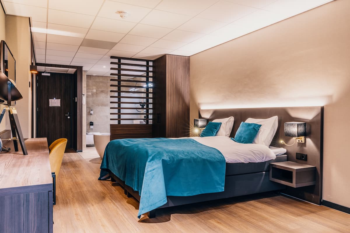 A modern hotel room with a large bed, a desk, and a chair. There is a nightstand with a lamp and a small shelf.