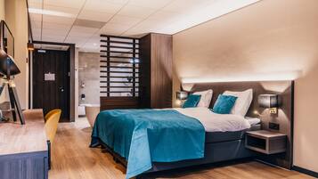 Una habitación de hotel moderna con una cama grande, un escritorio y una silla. Hay una mesita de noche con una lámpara y un estante pequeño.