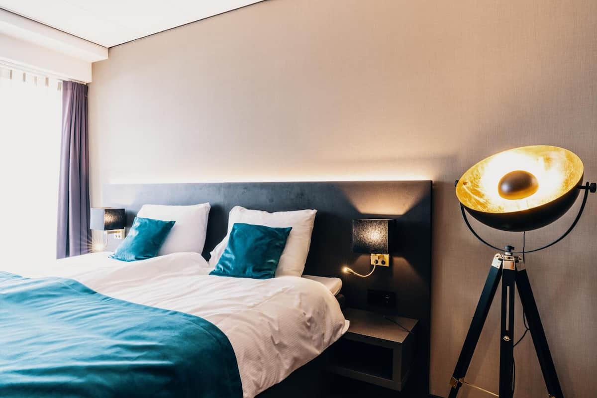 A neatly made bed with white and teal bedding, a bedside lamp, and a floor lamp on a tripod.