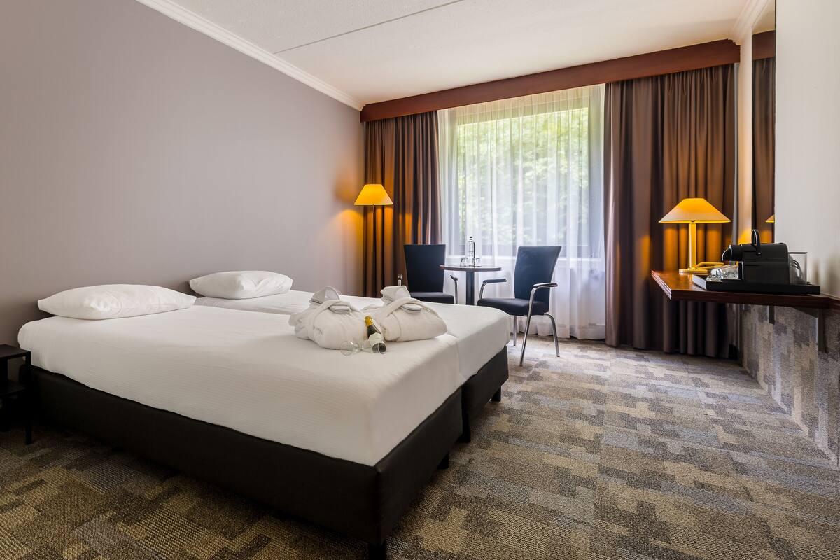 A hotel room with a large bed, two bedside tables, a desk with a chair, and a window with curtains.