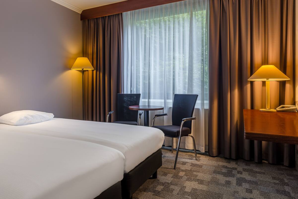 A hotel room with two beds, a small table, two chairs, a telephone, and a window with curtains.